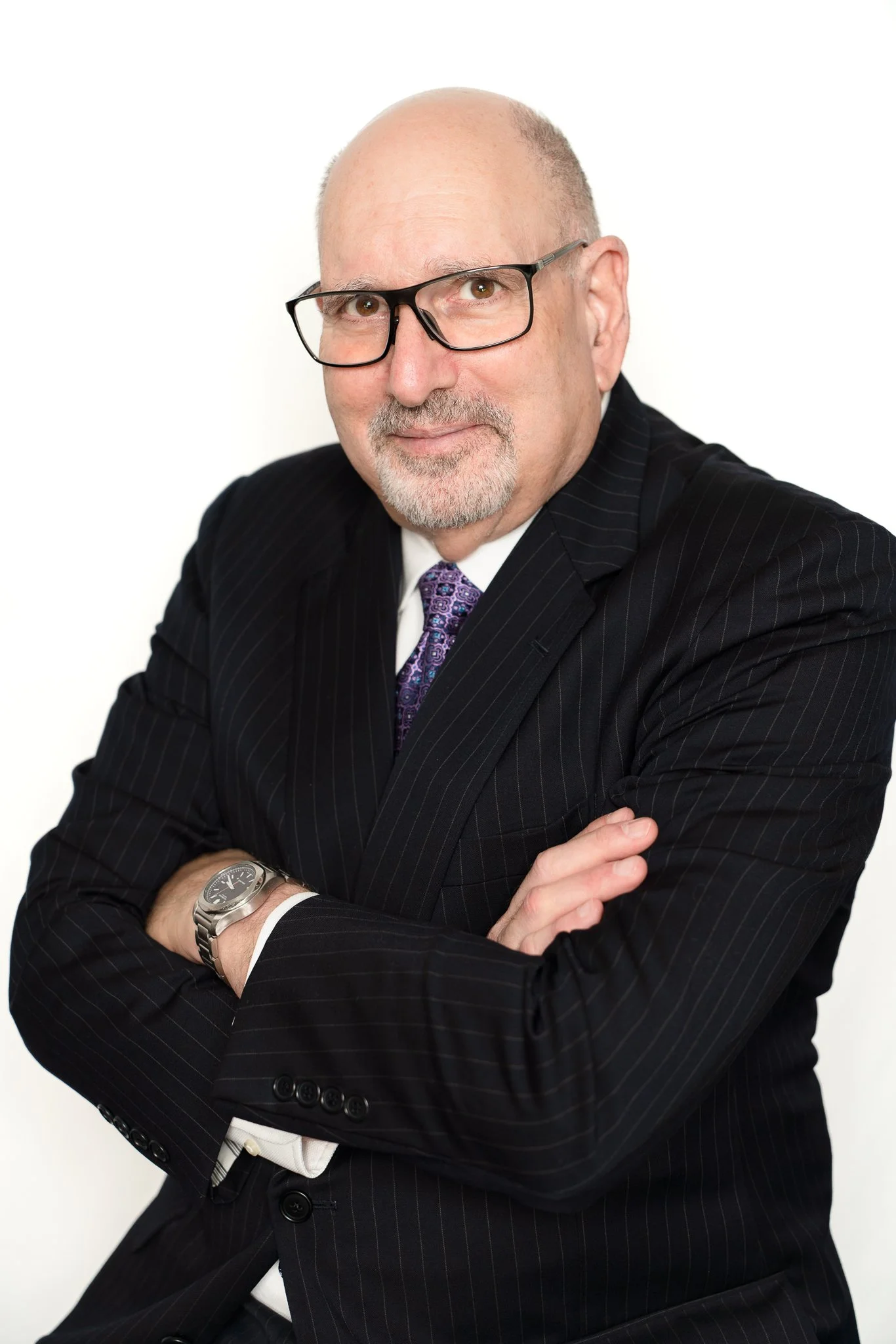 A middle-aged man in a pinstripe suit with glasses, white shirt, and purple tie, standing with arms crossed against a white background.