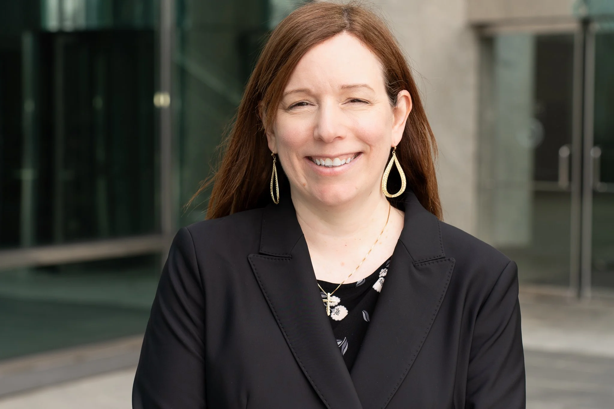 A woman with long brown hair, wearing a black blazer and a floral top, smiling outdoors with a modern glass building in the background.