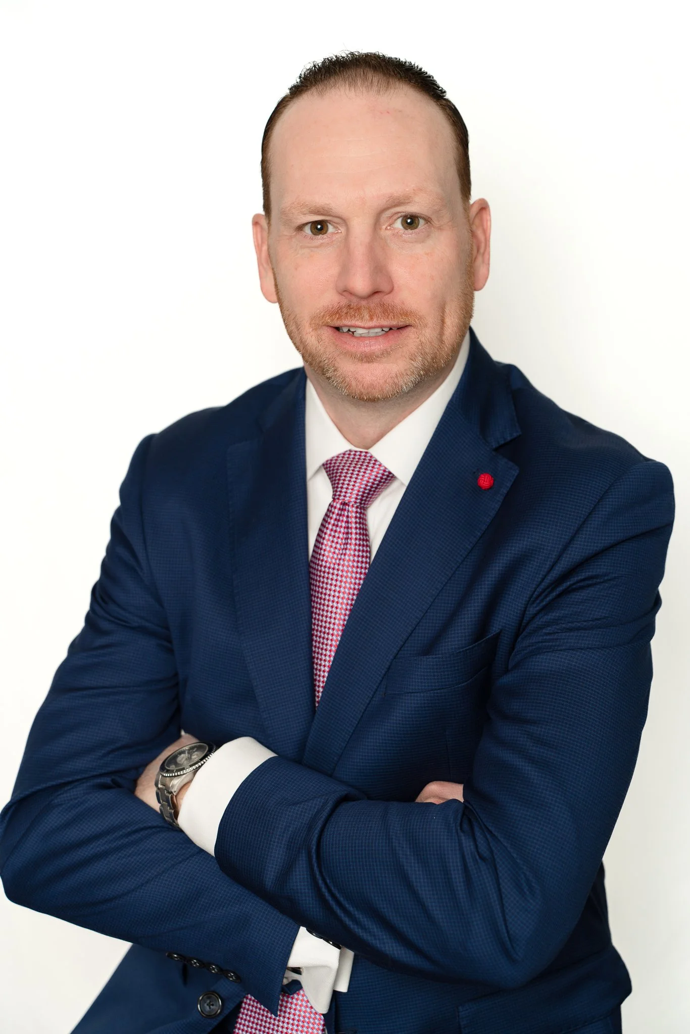 A man in a navy blue suit, white shirt, red and white checkered tie, and silver watch, standing against a white background with arms crossed and smiling.