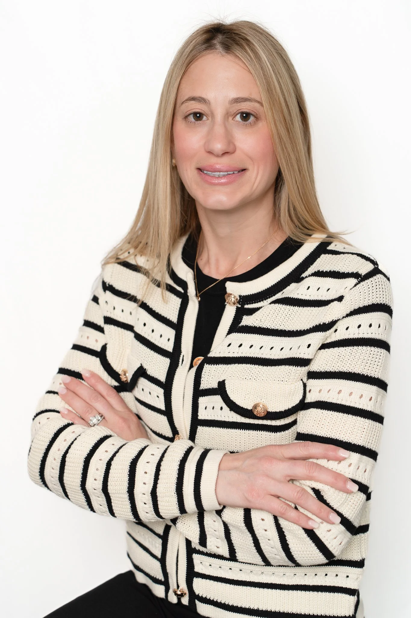 A woman with blonde hair in a striped cream and black sweater crossed arms, smiling slightly, standing against a white background.