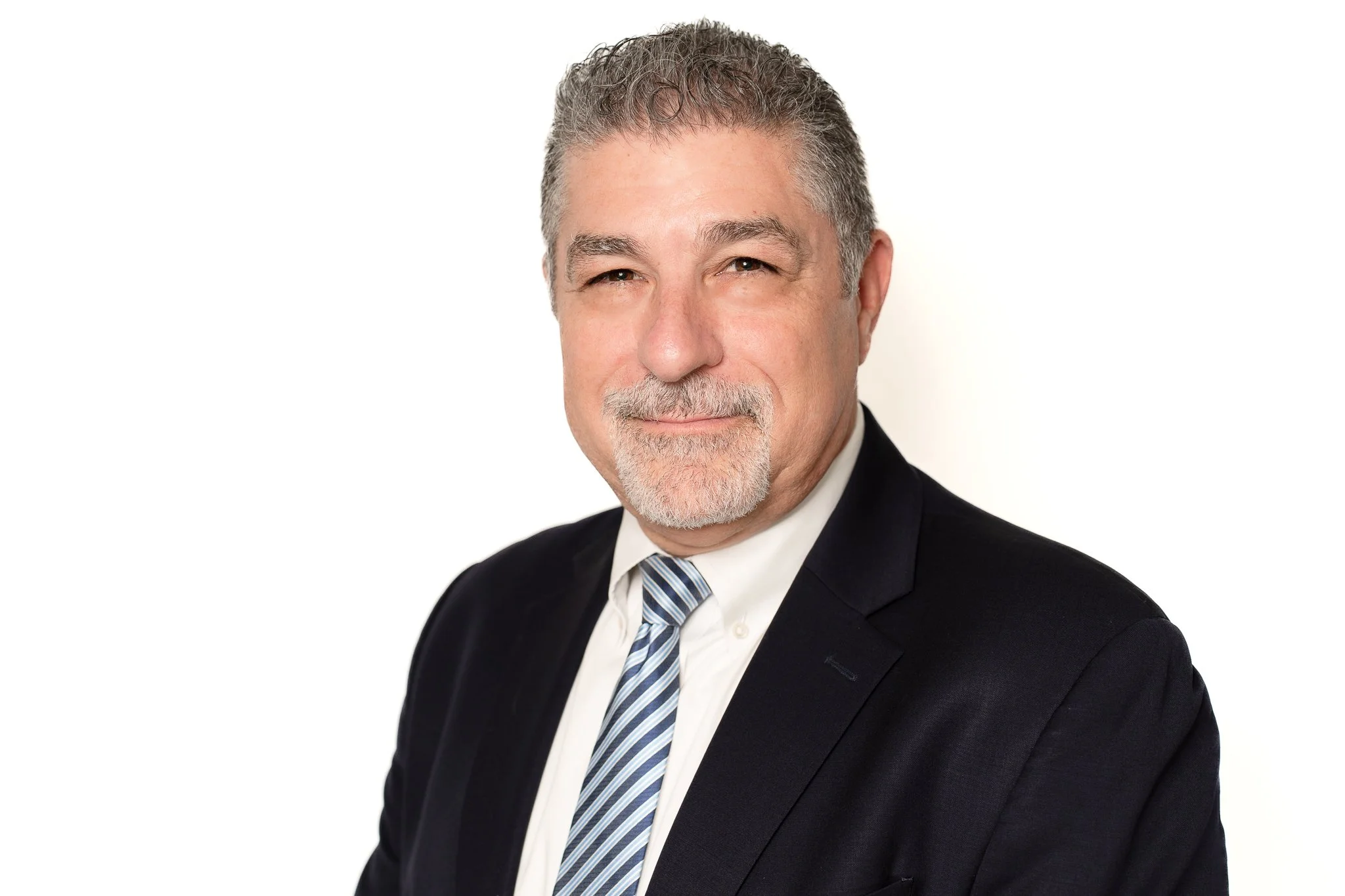 A middle-aged man with gray hair and a goatee, dressed in a black suit, white shirt, and striped tie, smiling against a plain white background.