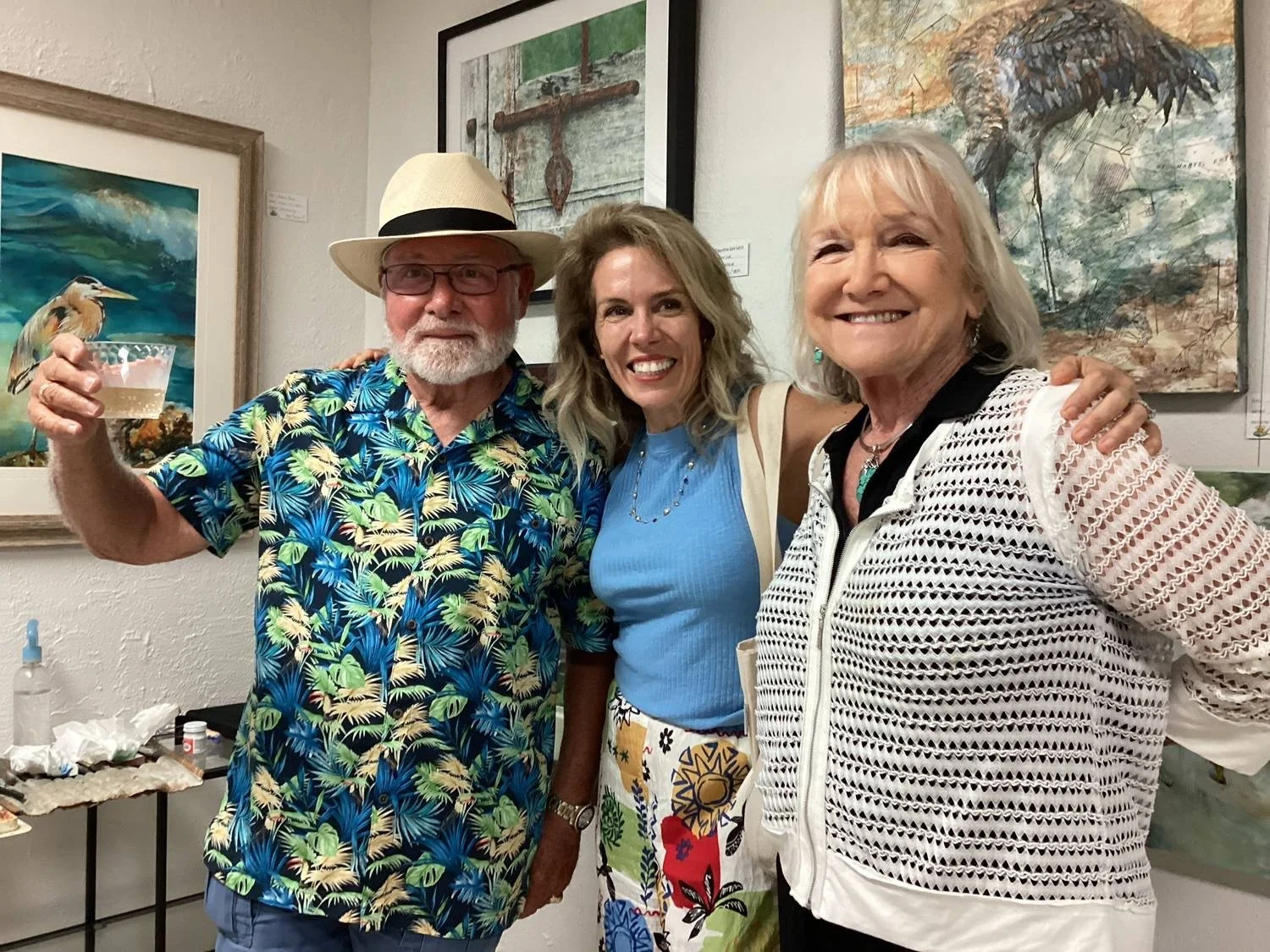 Three people, two women and one man, are smiling and posing together at an art gallery. The man is wearing a floral shirt and a white hat with a black band, holding a drink. The woman in the middle is wearing a blue top and a colorful skirt. The woman on the right is wearing a white jacket with black accents.