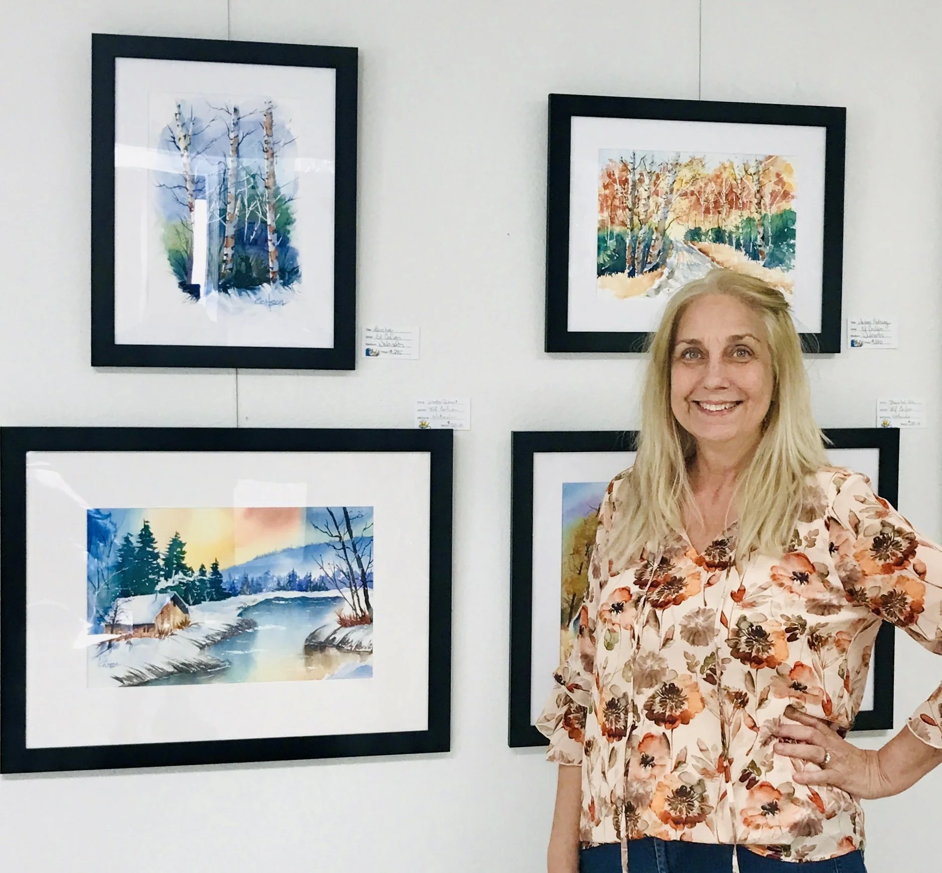A woman with blonde hair smiling at an art gallery, standing in front of four framed watercolor landscape paintings depicting trees, a river, and a cabin.