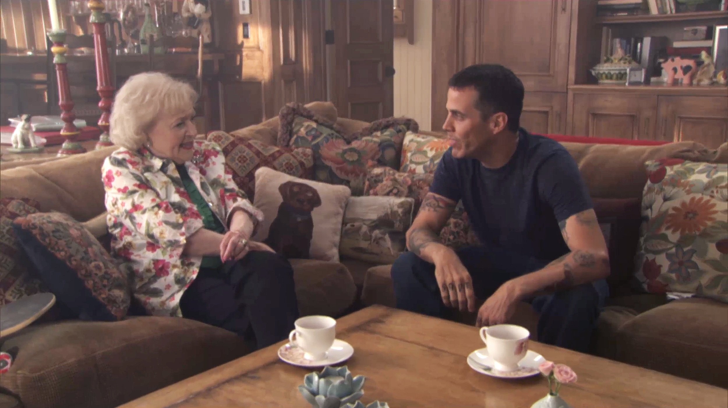 Two people sitting on a couch in a cozy living room with colorful cushions and a wooden table with teacups.