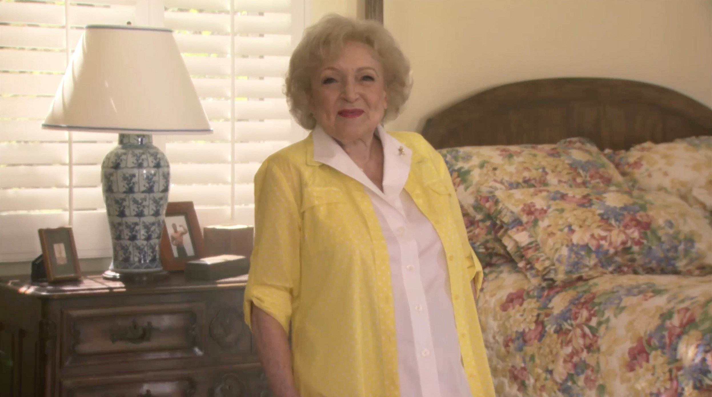Elderly woman in a yellow shirt standing in a bedroom with floral bedding, a wooden headboard, and a nightstand with a decorative blue and white lamp.