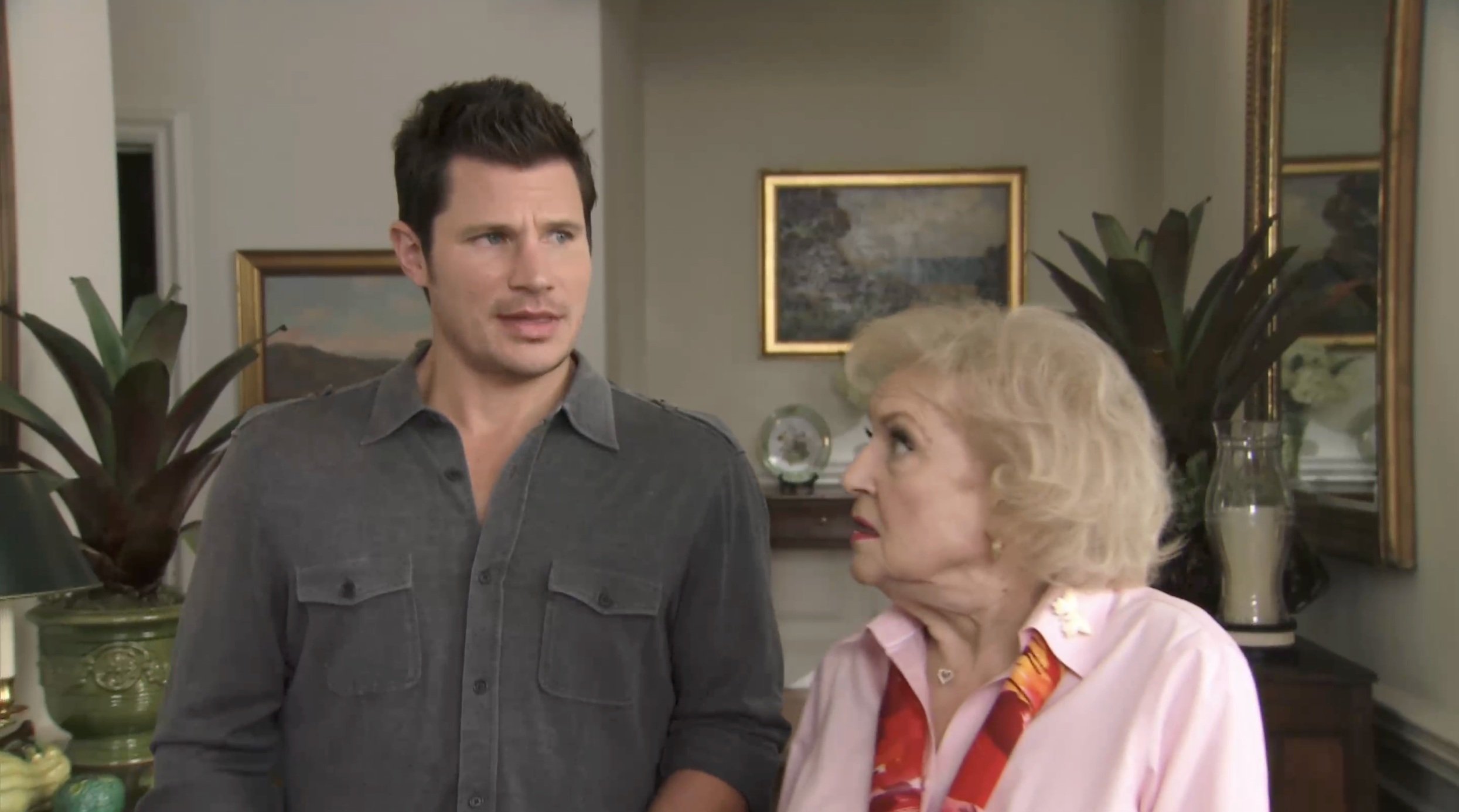 A man and an elderly woman standing indoors, looking towards each other, with framed paintings and a plant in the background.
