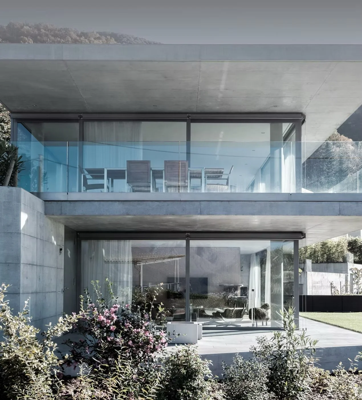 Casa moderna con pareti di vetro e balcone con tavolo e sedie, giardino con piante e fiori colorati.