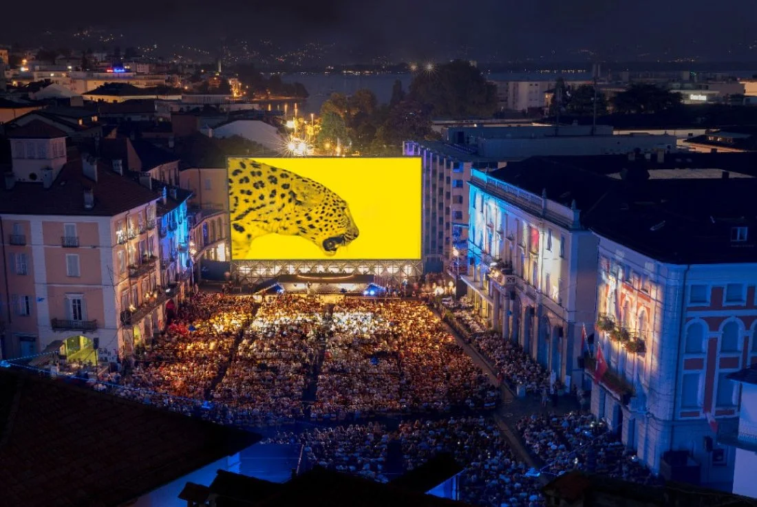 Un grande schermo mostra un felino giallo con macchie nere in una piazza affollata di persone, con edifici illuminati sullo sfondo, durante la notte.