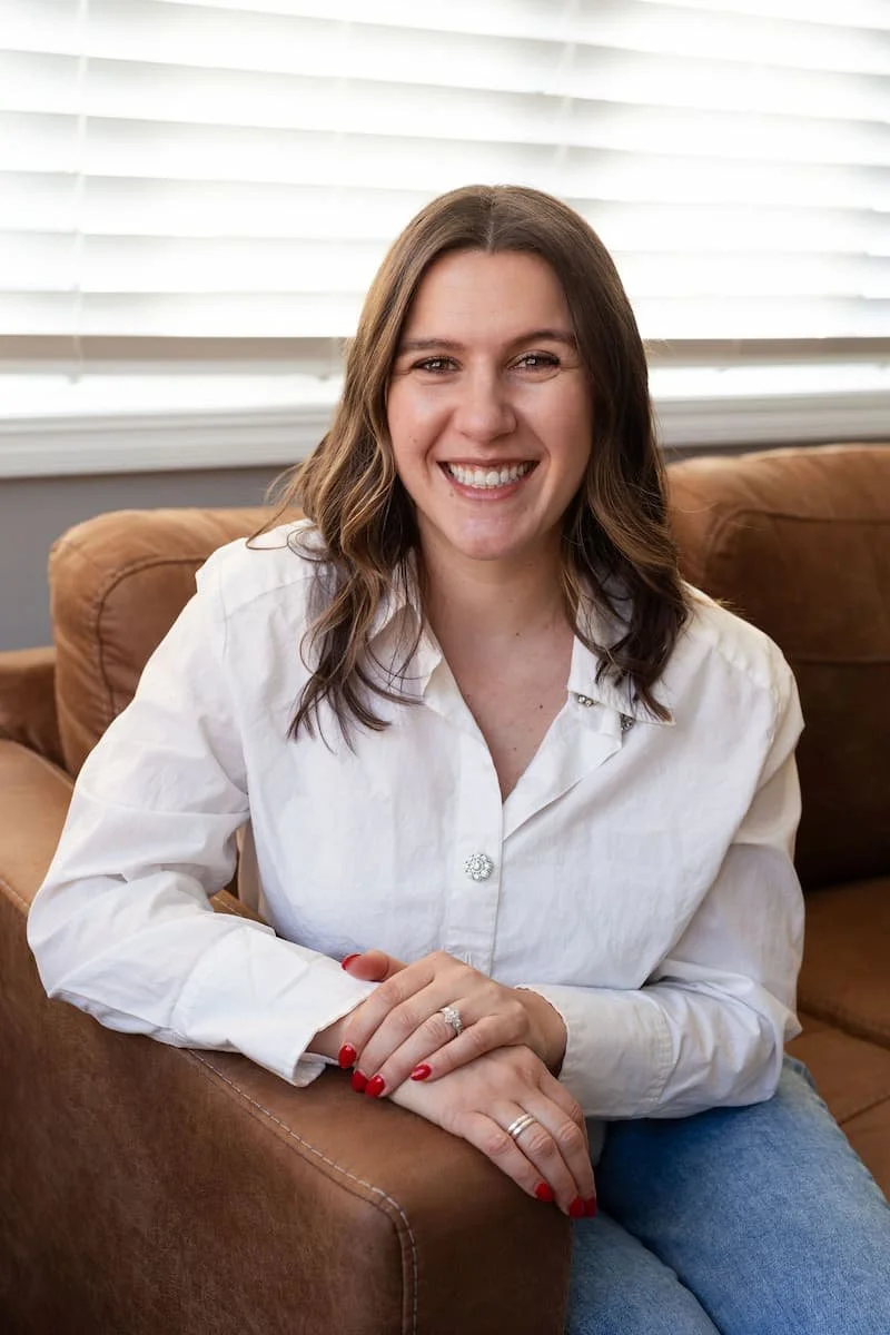 A woman with shoulder-length brown hair, smiling and wearing a white blouse, sitting on a brown couch in front of a window with white blinds.