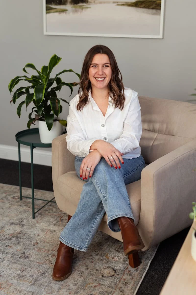 A woman with shoulder-length brown hair, wearing a white button-up shirt and blue jeans, sitting on a beige armchair in a living room, smiling with her hands folded on her lap. Behind her is a large green houseplant on a small round table, and a framed landscape photograph on the wall.