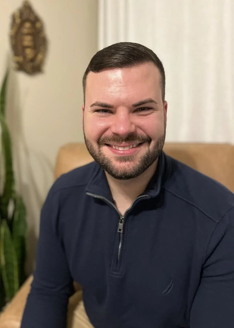 A smiling man with short dark hair and a beard, wearing a navy blue zip-up sweater, sitting indoors with a beige chair, a potted plant, and a decorative wall hanging in the background.