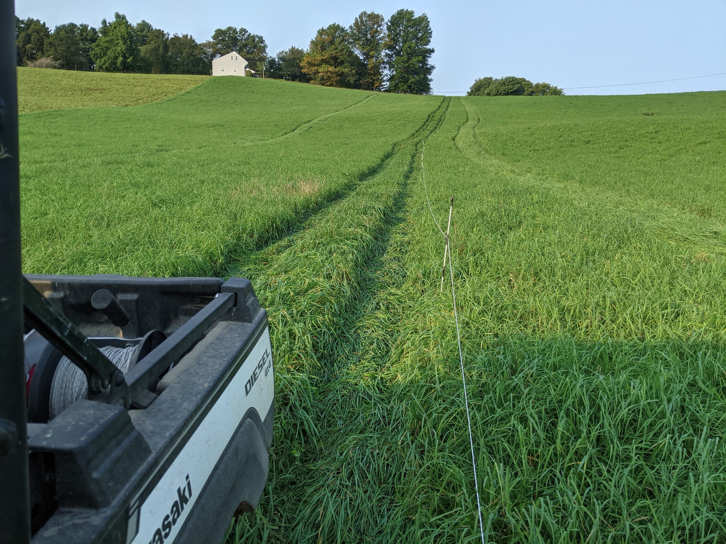 Green grassy field with a white house on a hill in the background, tire tracks and a measuring tape in the foreground, blue sky, trees at the top of the hill.