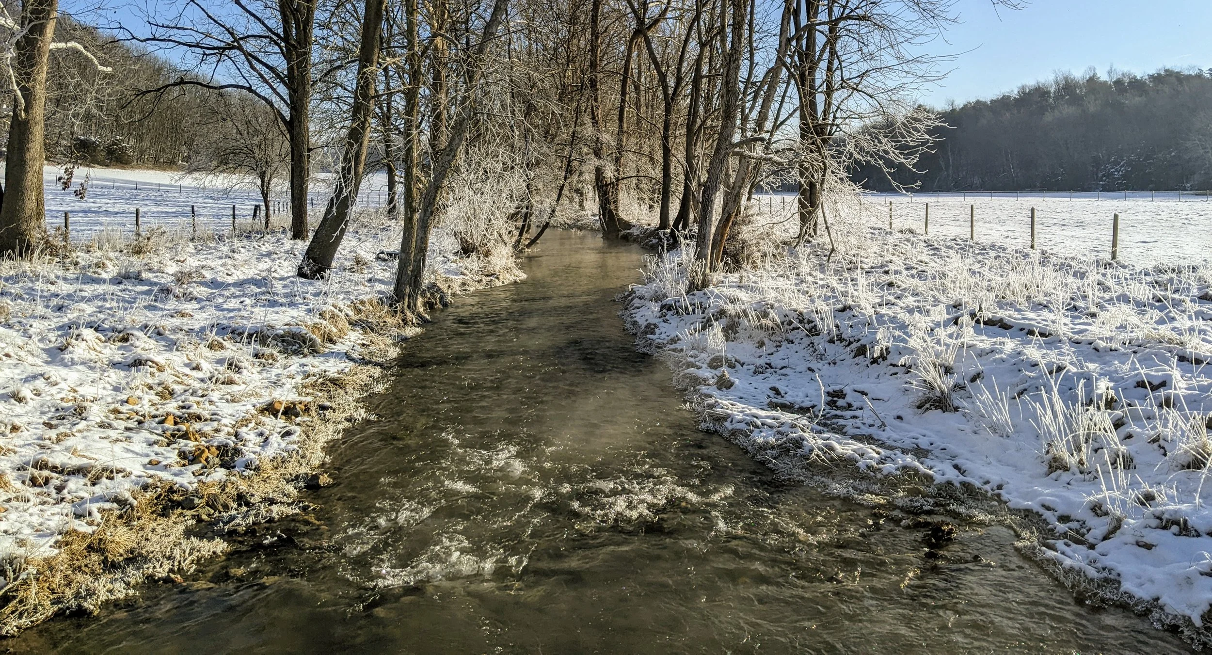 Snow-covered landscape with a small creek, trees with frosty branches, snowy fields, and fencing under a clear blue sky.