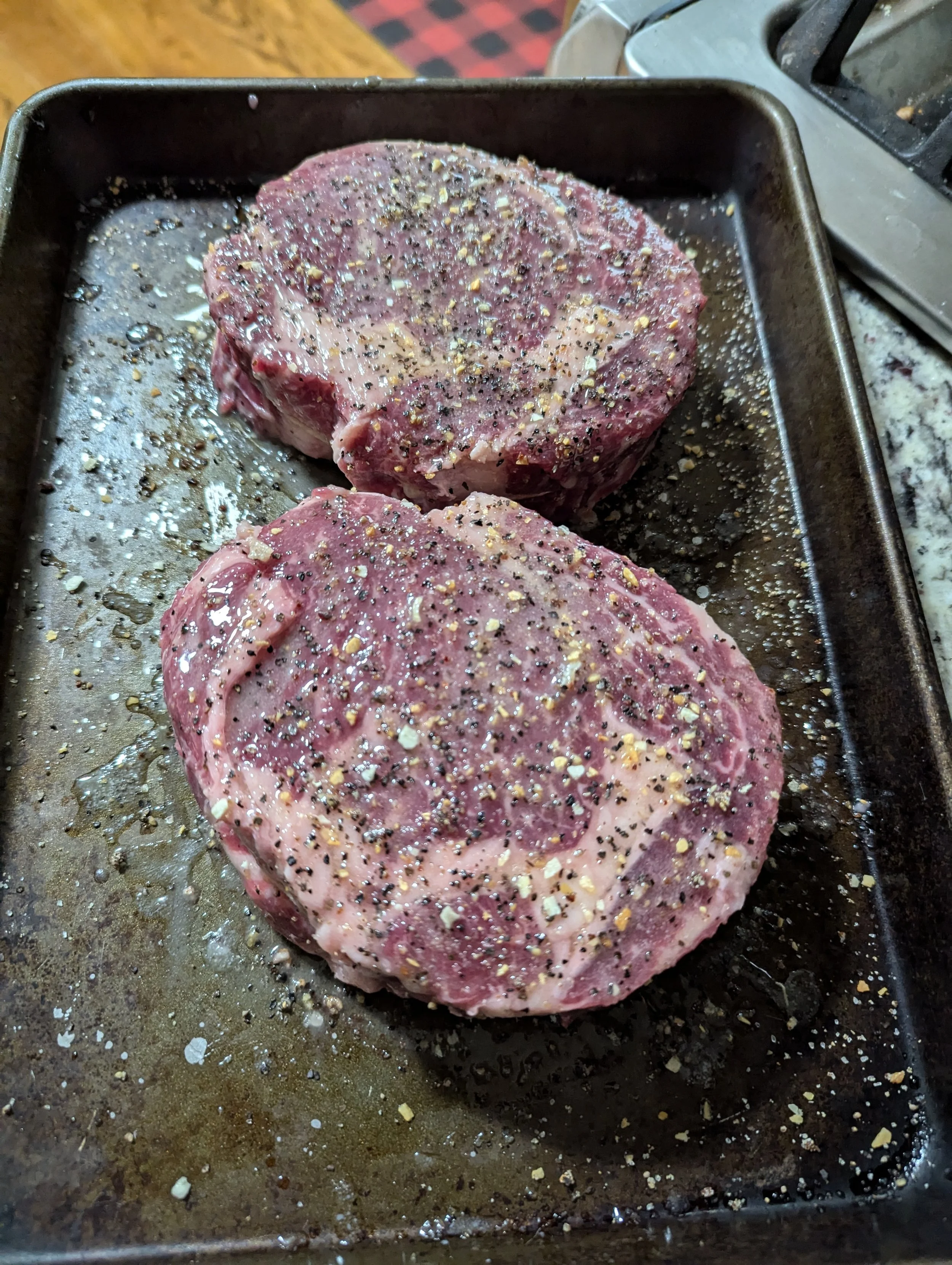 Two raw steaks seasoned with salt, black pepper, and herbs on a baking sheet.