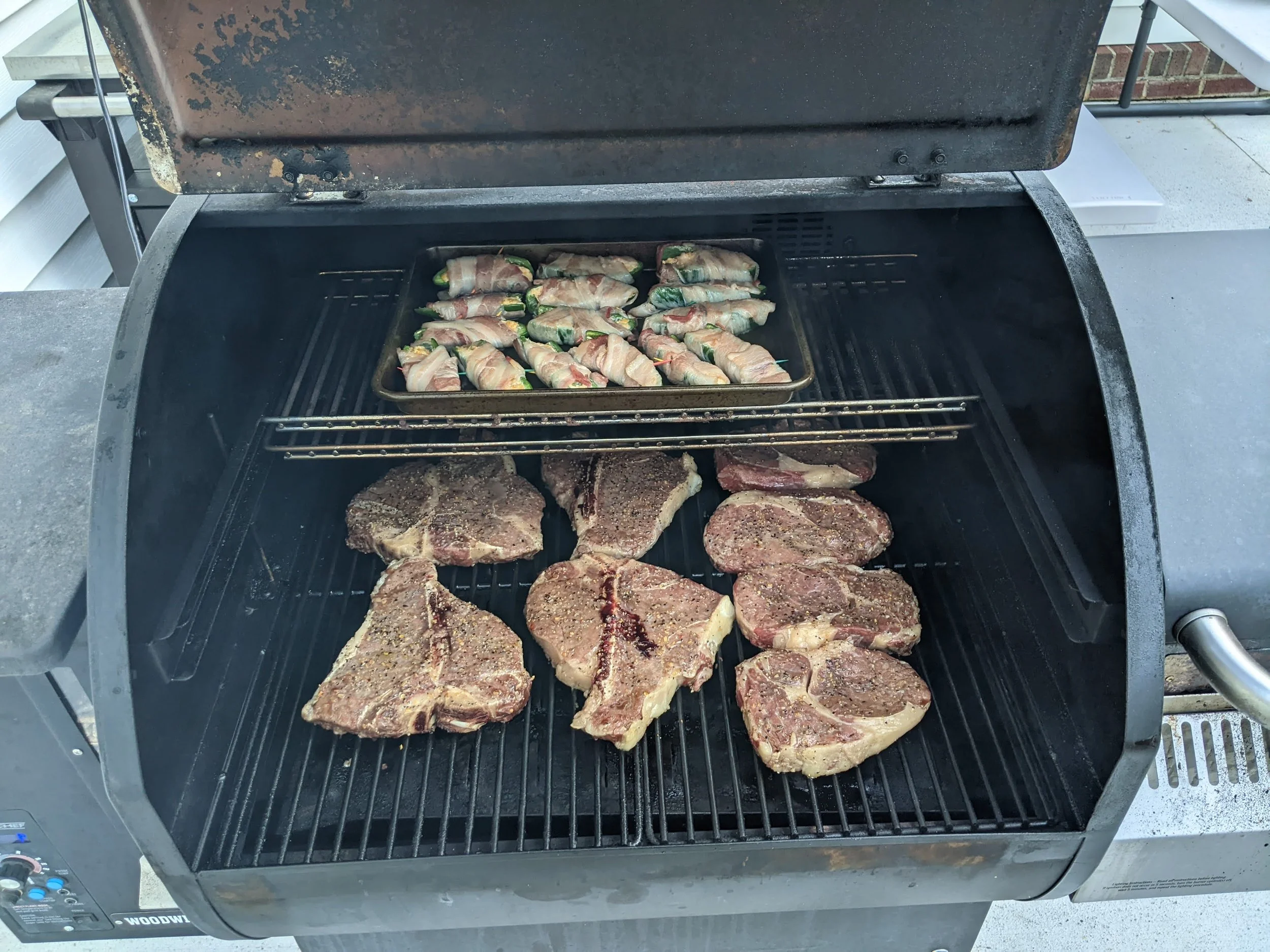 A pellet grill outdoor barbecue with raw seasoned steaks and bacon-wrapped vegetables cooking on the grill grate.