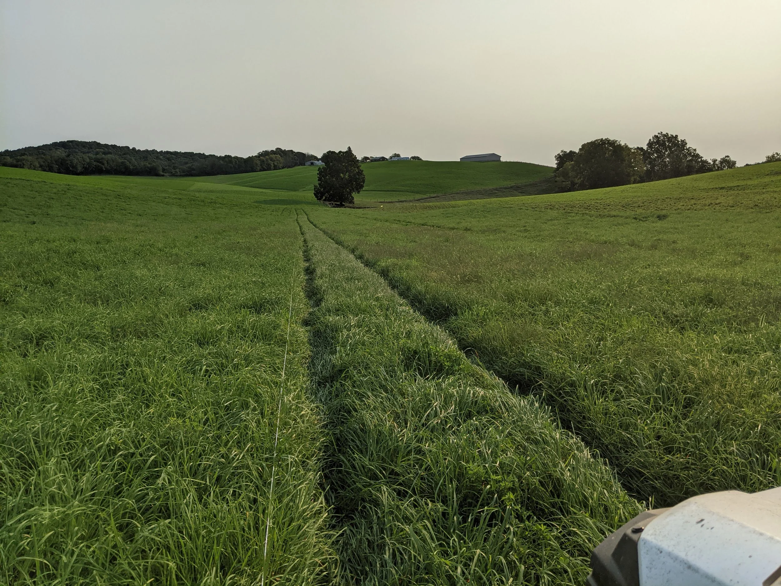 Green rolling hills with green grass, scattered trees, and farm buildings in the distance under a clear sky. Part of a vehicle or equipment visible in the bottom right corner.