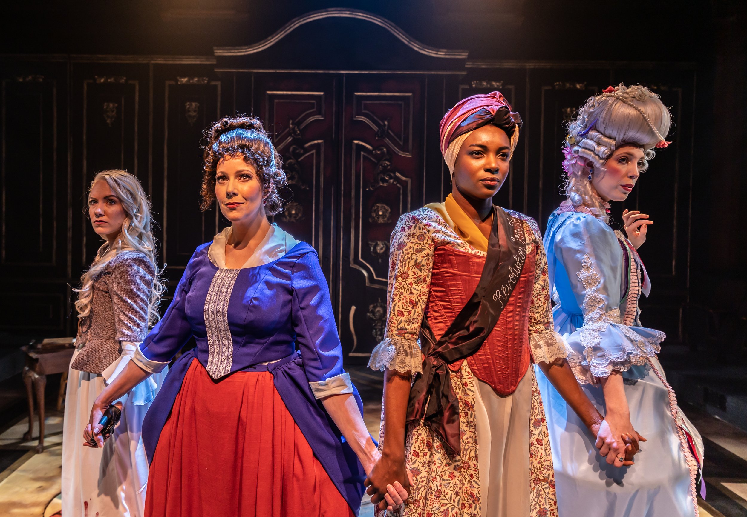 Four women in historical costumes are standing in a line, holding hands, on a stage with a dark, ornate backdrop.