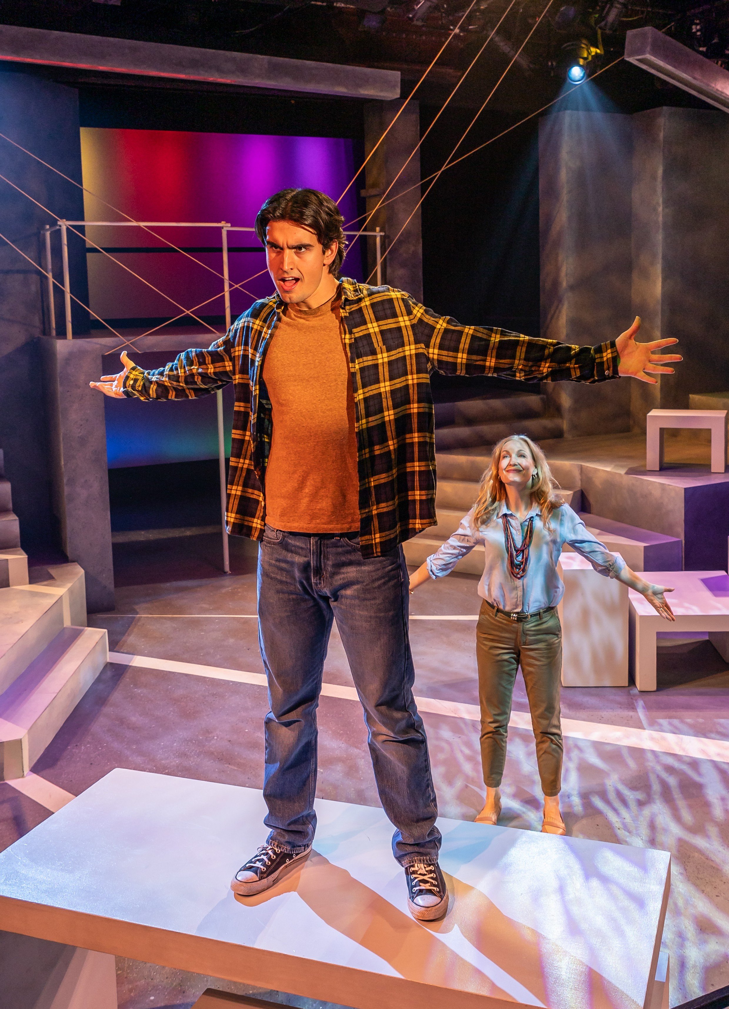 Two actors performing on stage with one standing on a white platform in the foreground and the other behind, with dramatic lighting and staged set design.