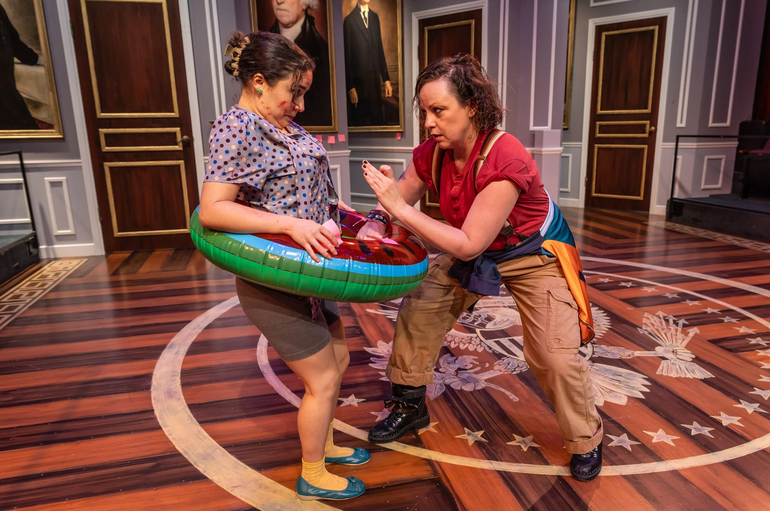 Two women in a staged setting resembling a government chamber, one with a bloody face and injury, wearing a polka dot shirt with a float ring around her waist, and the other woman with short dark hair, wearing a red shirt and khaki pants, holding her and gesturing as if in a confrontation.