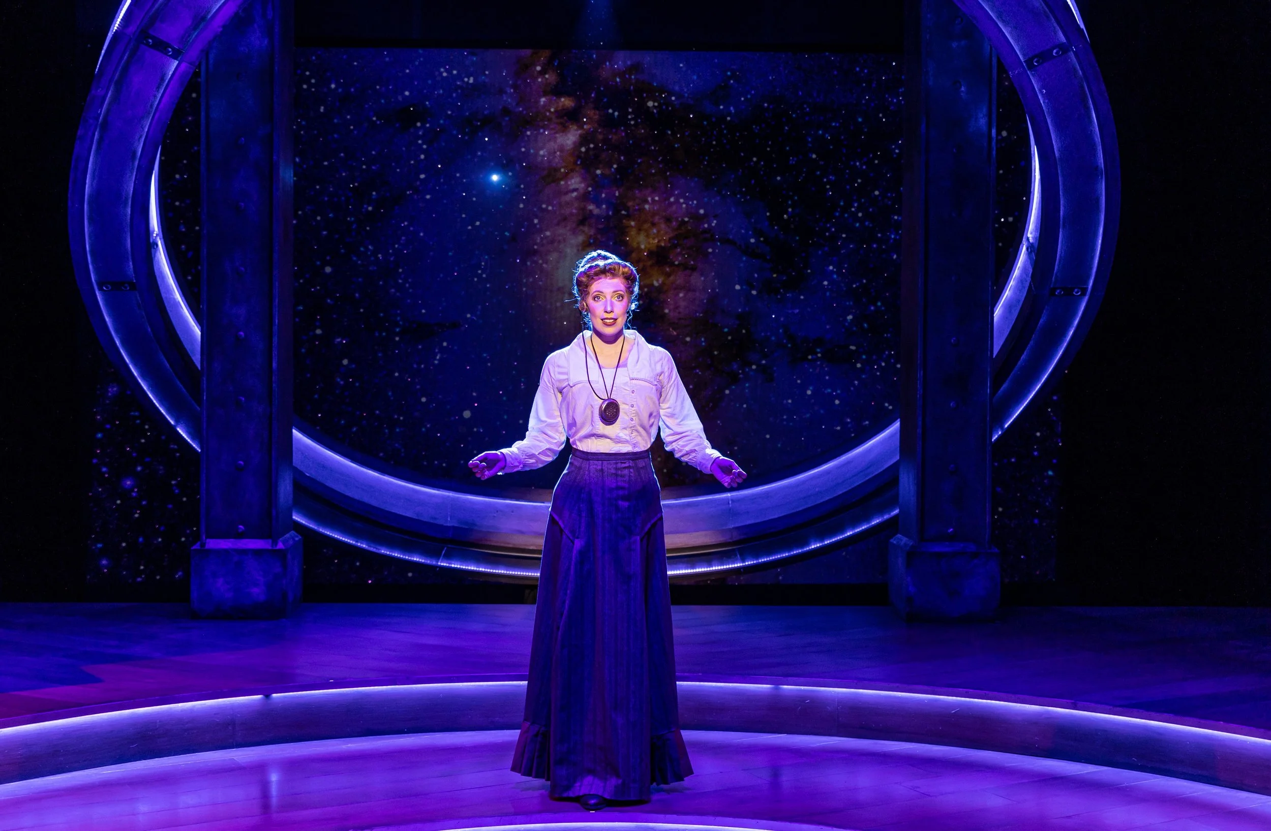 A woman standing on a stage with a backdrop of galaxy stars, framed by a futuristic archway, illuminated with purple and blue lighting.