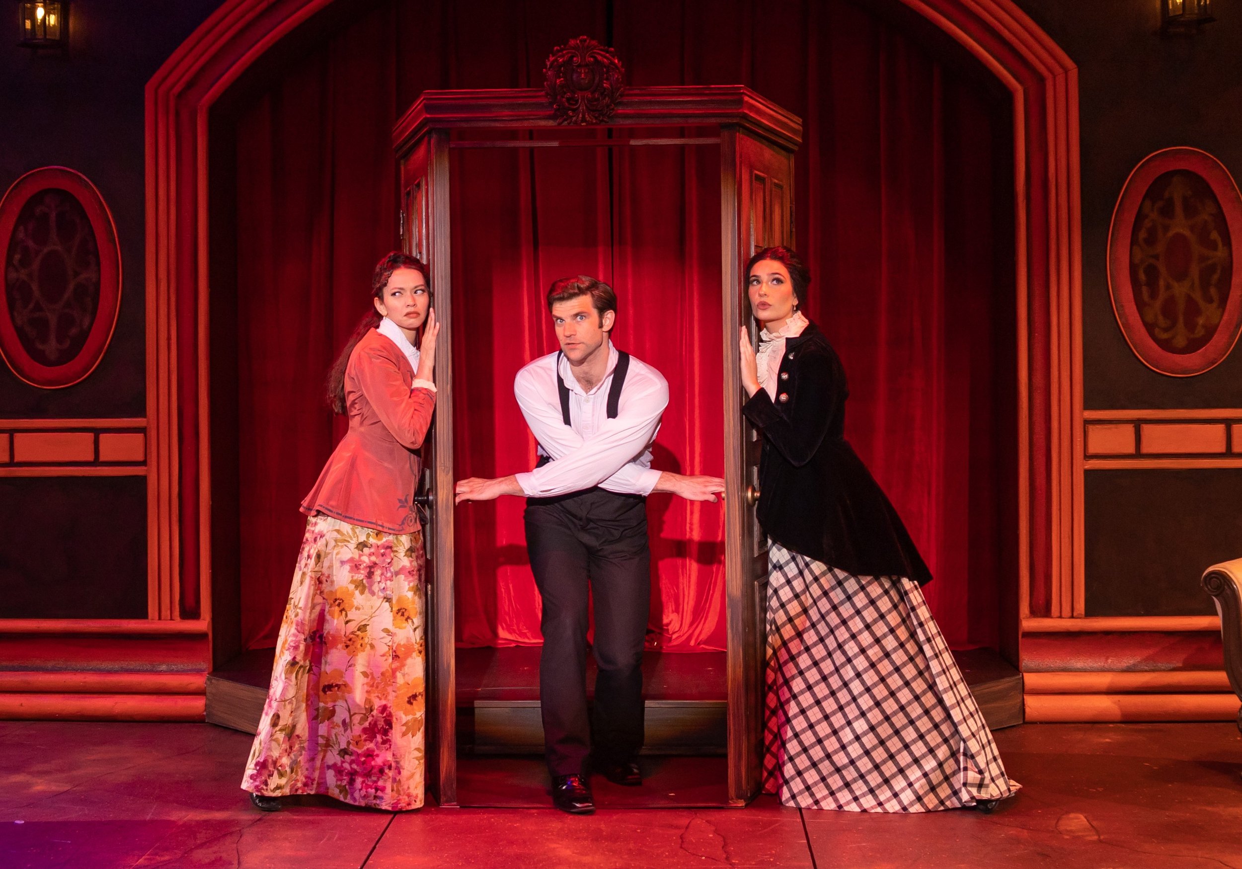 Three actors on stage in period costumes perform in a theater with red curtains and wooden decor. Two women stand on either side of a man who is leaning through an open door.
