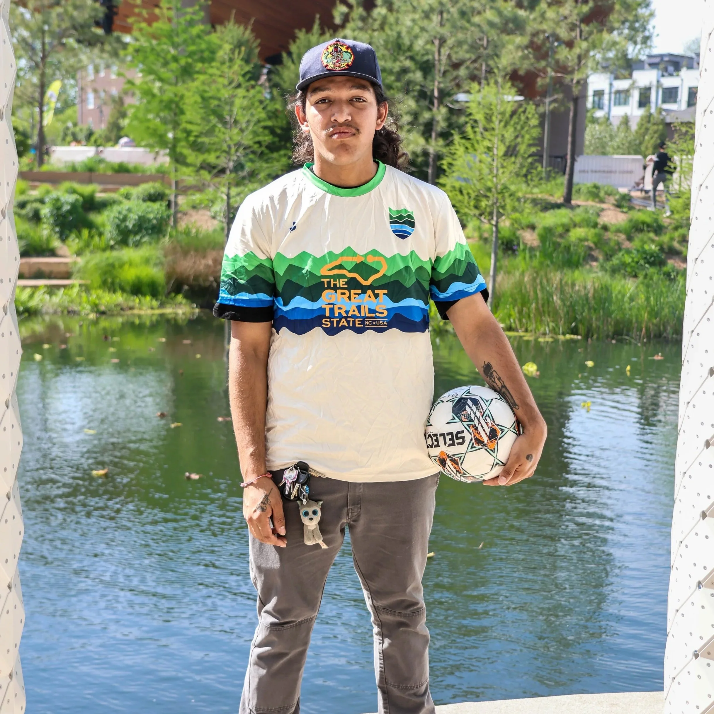 Young man standing outdoors by a pond, holding a soccer ball, wearing a themed t-shirt, baseball cap, and gray pants, with green trees and buildings in the background.