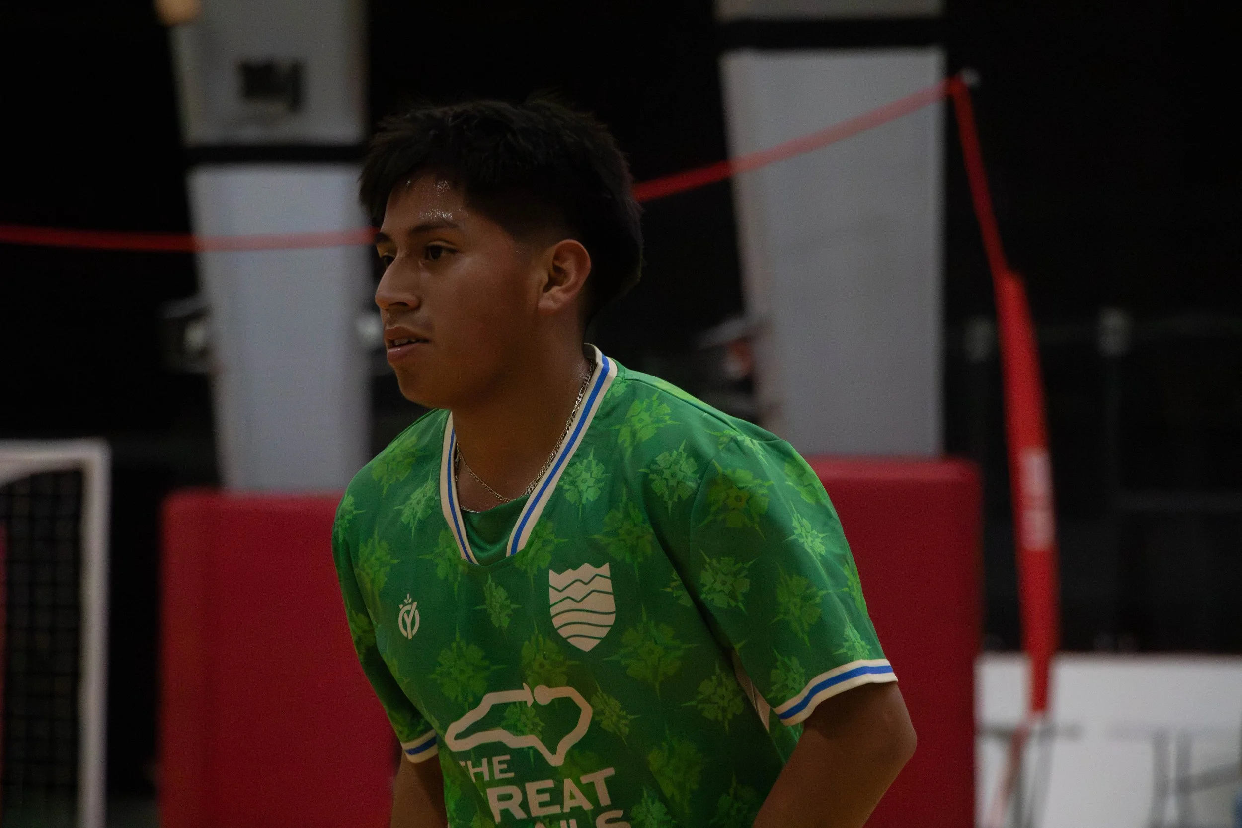 Young male athlete with dark hair, wearing a green sports jersey with white and blue accents, standing indoors with a red barrier and equipment in the background.