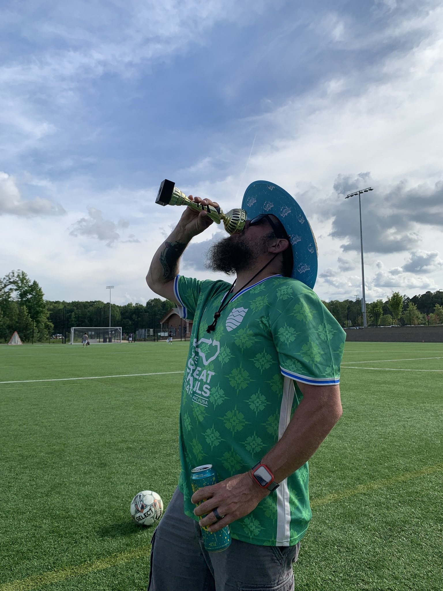 A man with a beard and tattoos wearing a green sports jersey, wide-brimmed hat, and smartwatch, drinking from a trophy cup on a soccer field. There are soccer goals and players in the background, with a blue sky and scattered clouds overhead.