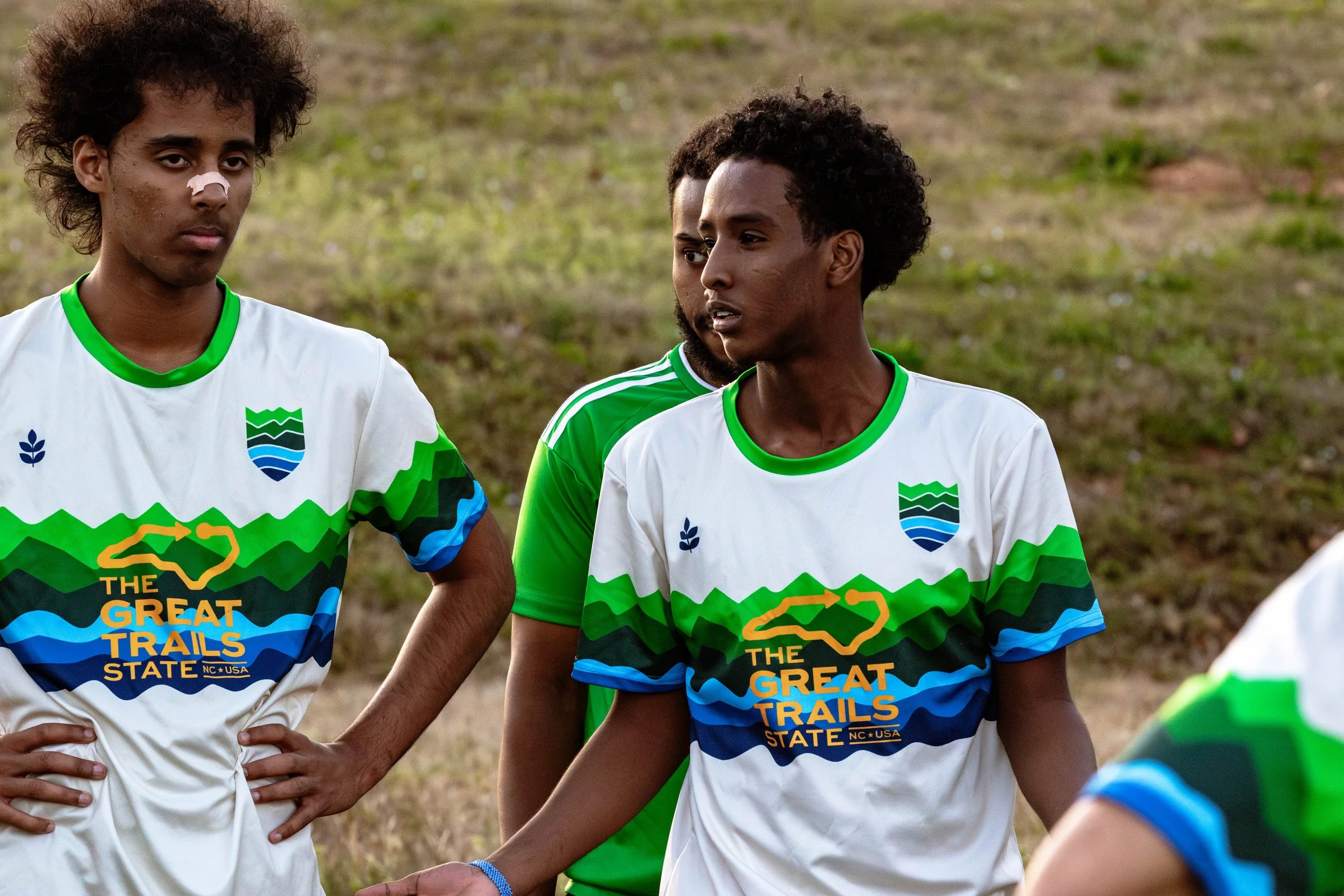 Three young male athletes in sports jerseys standing outdoors on a grassy field, engaged in a discussion during a game or practice.