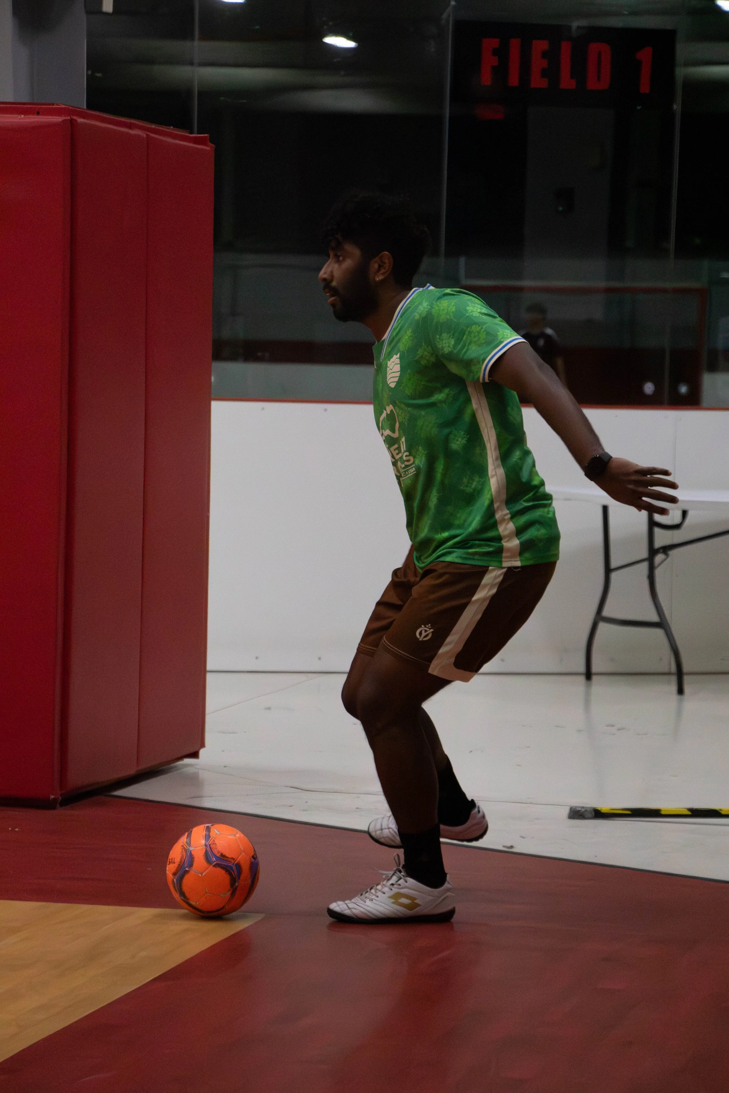 A man in a green sports jersey and brown shorts preparing to kick a soccer ball on an indoor court.
