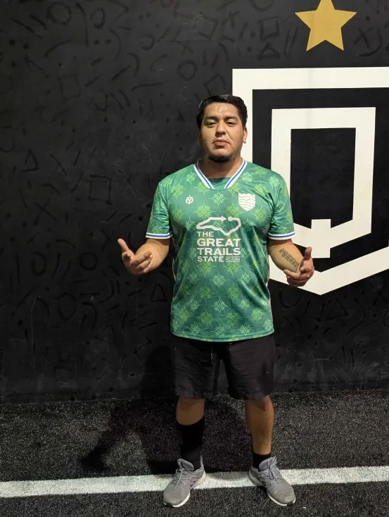 Man posing in front of a black wall with a Twitch logo; wearing a green sports jersey and black shorts.