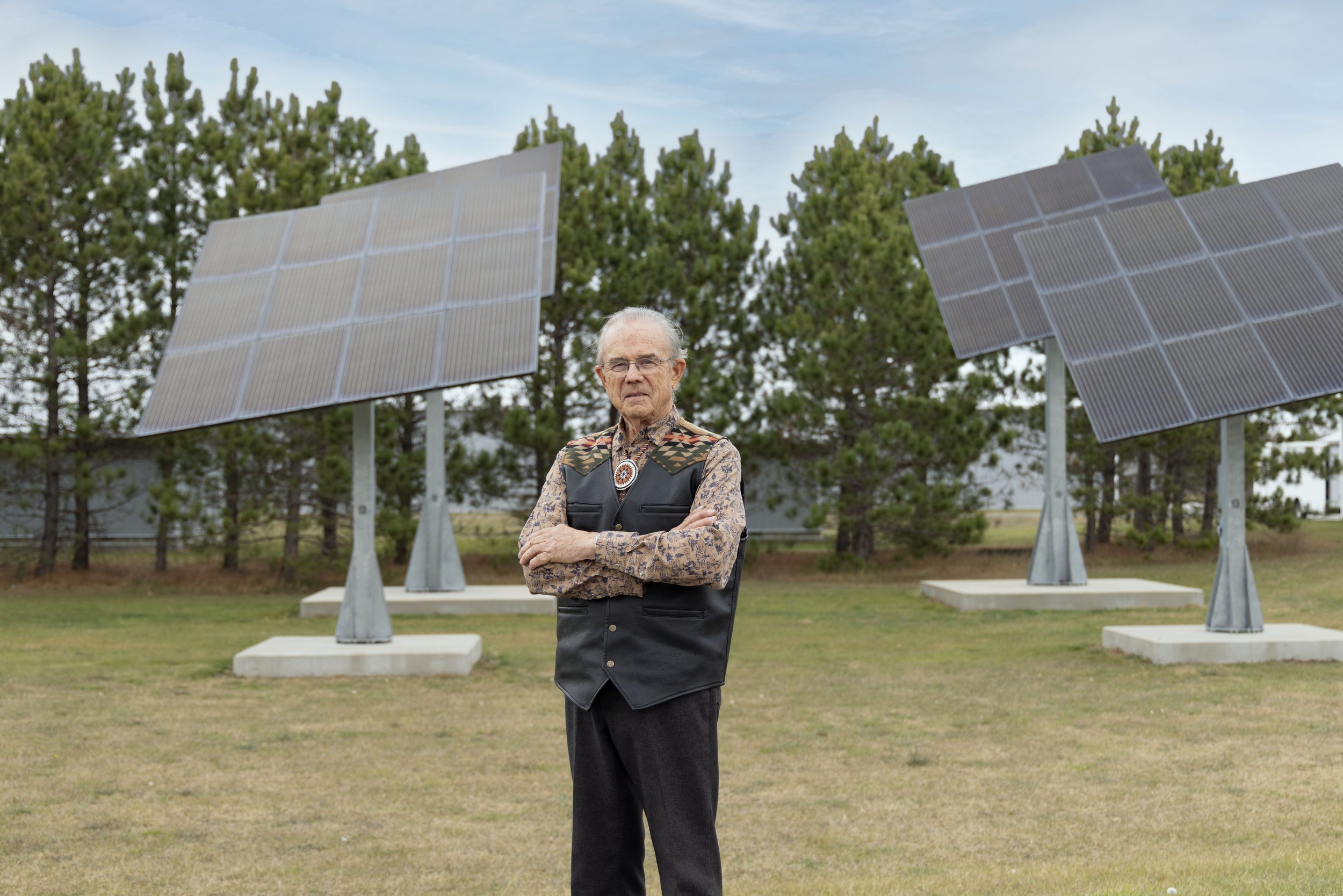 An elderly man with gray hair and glasses standing with arms crossed in front of solar panels on a grassy area with trees in the background.