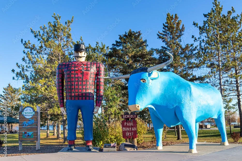 Large artistic statues of a human figure dressed in a red and black checkered shirt and blue pants, and a big blue bull with white horns, located outdoors in a park setting with trees and a clear blue sky.