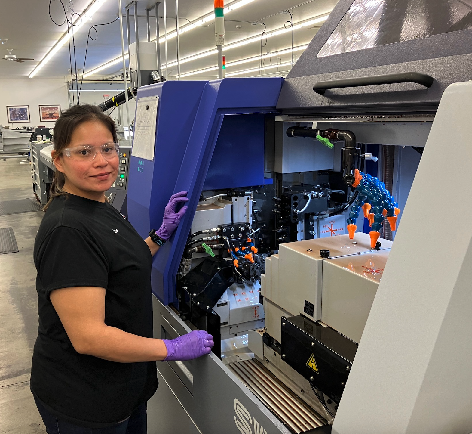 A woman wearing safety glasses, purple gloves, and a black T-shirt standing beside a piece of advanced scientific machinery in a laboratory or manufacturing environment.