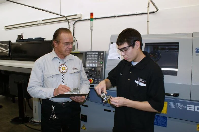 Two men working in a manufacturing or machine shop; one older man taking notes on a clipboard, the younger man inspecting a metal part.
