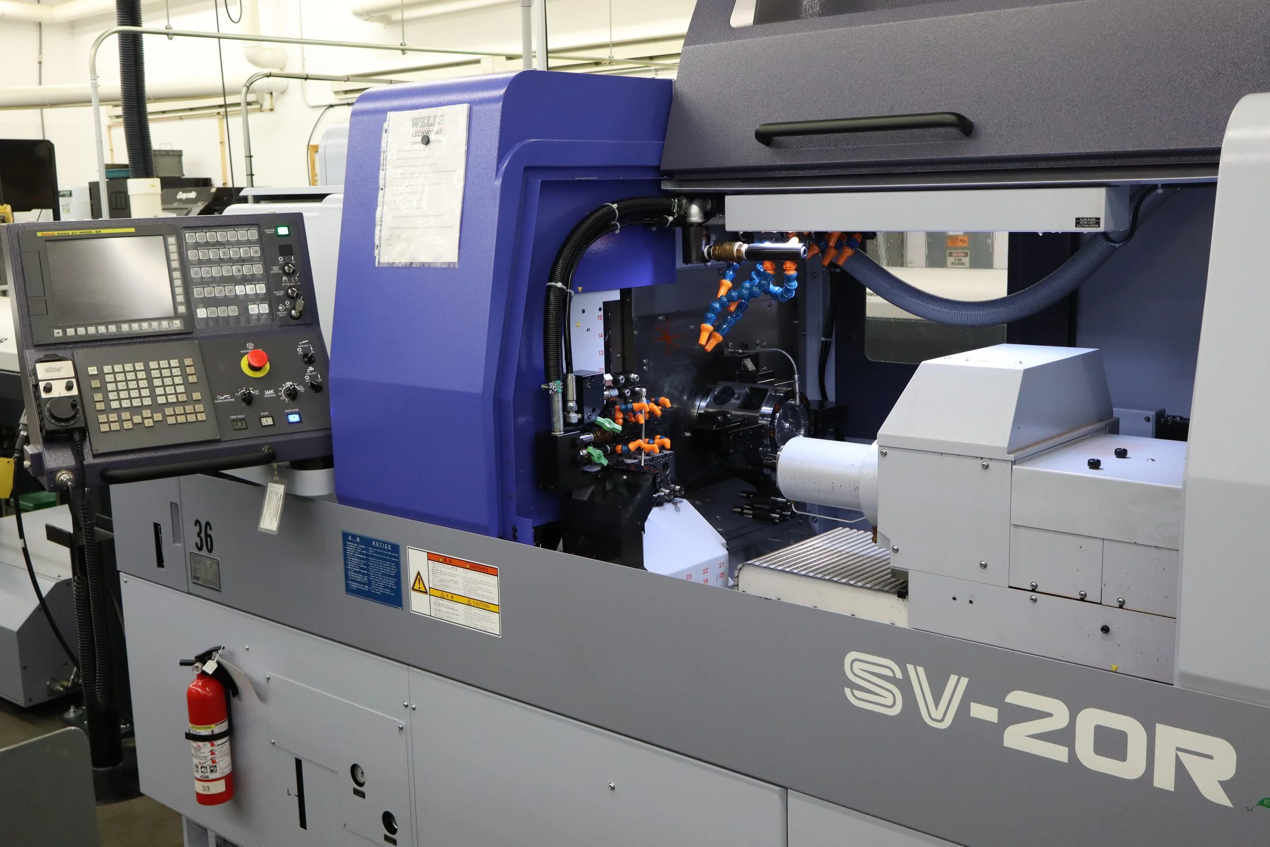 Industrial CNC machine with control panel on the left, part of the machine's interior visible with various hoses and tools, in a manufacturing facility.