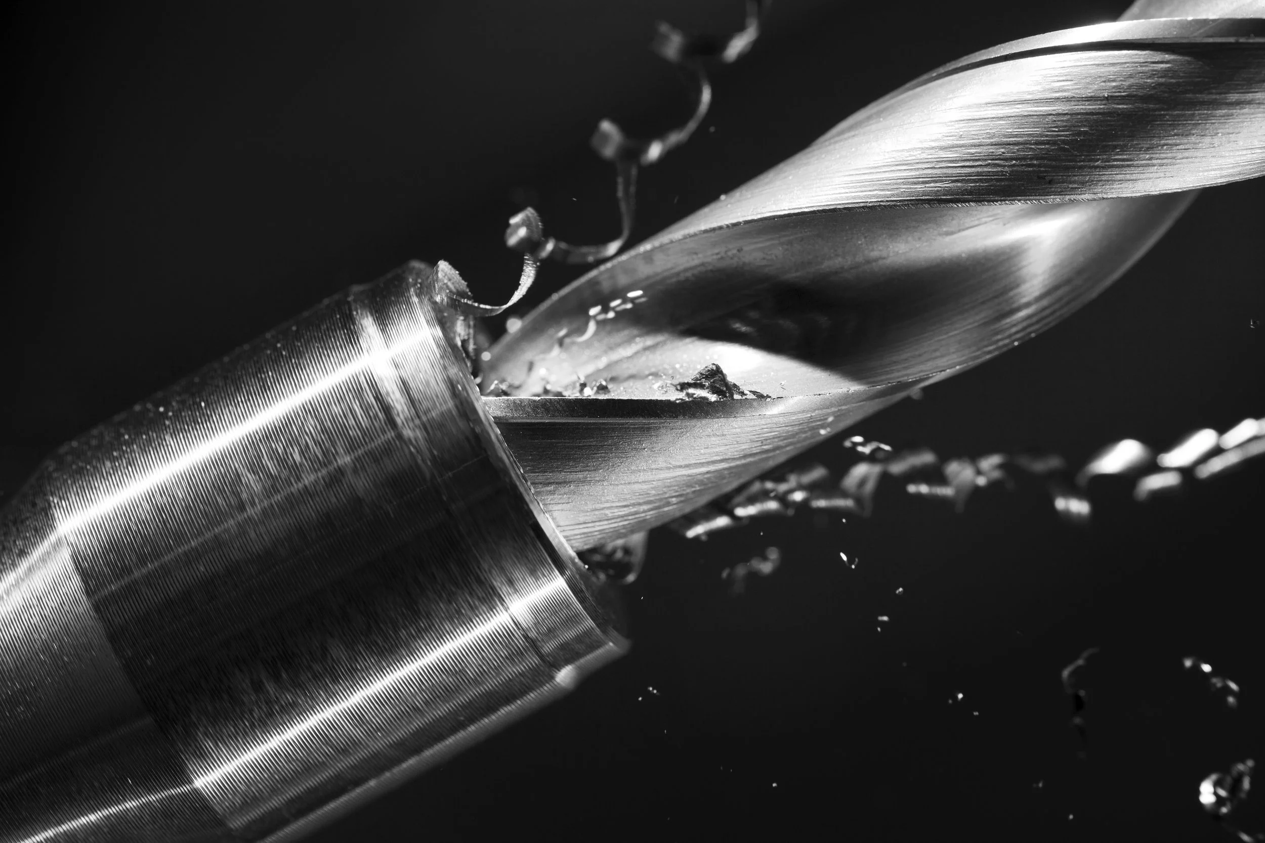 Close-up black and white image of a metal drill bit cutting into a cylindrical metal object, with metal shavings falling around.