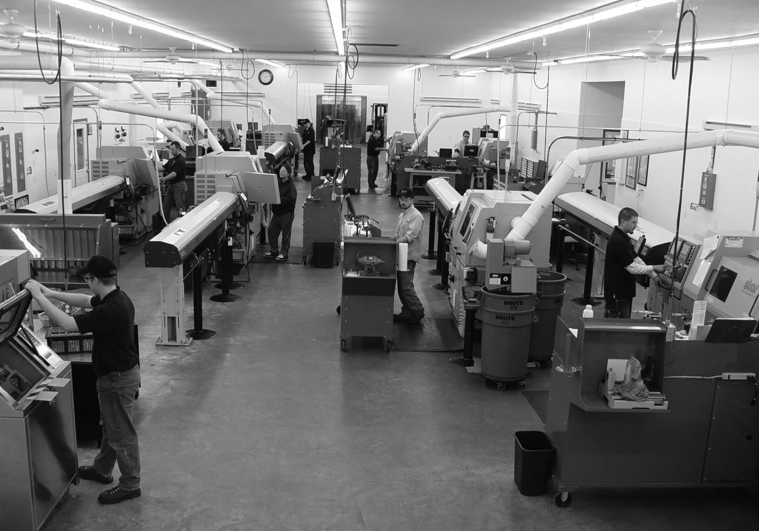 People working with machinery inside a factory or laboratory, with large equipment and workstations across the room, in black and white.