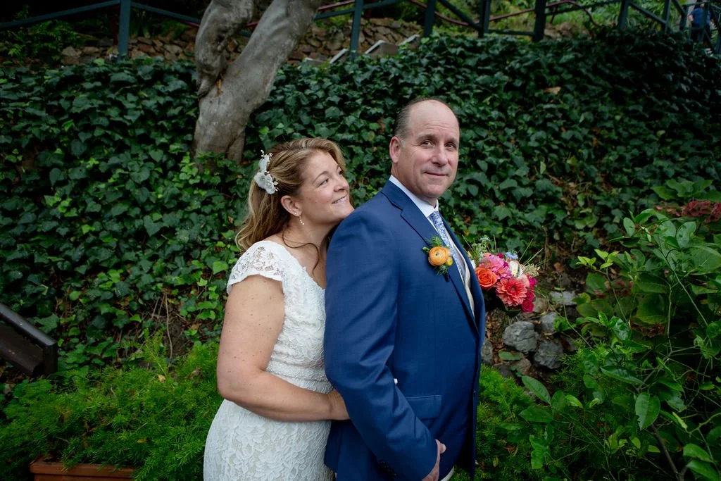 A bride in a white dress lovingly embraces a groom in a blue suit holding a colorful bouquet, with greenery and ivy in the background.