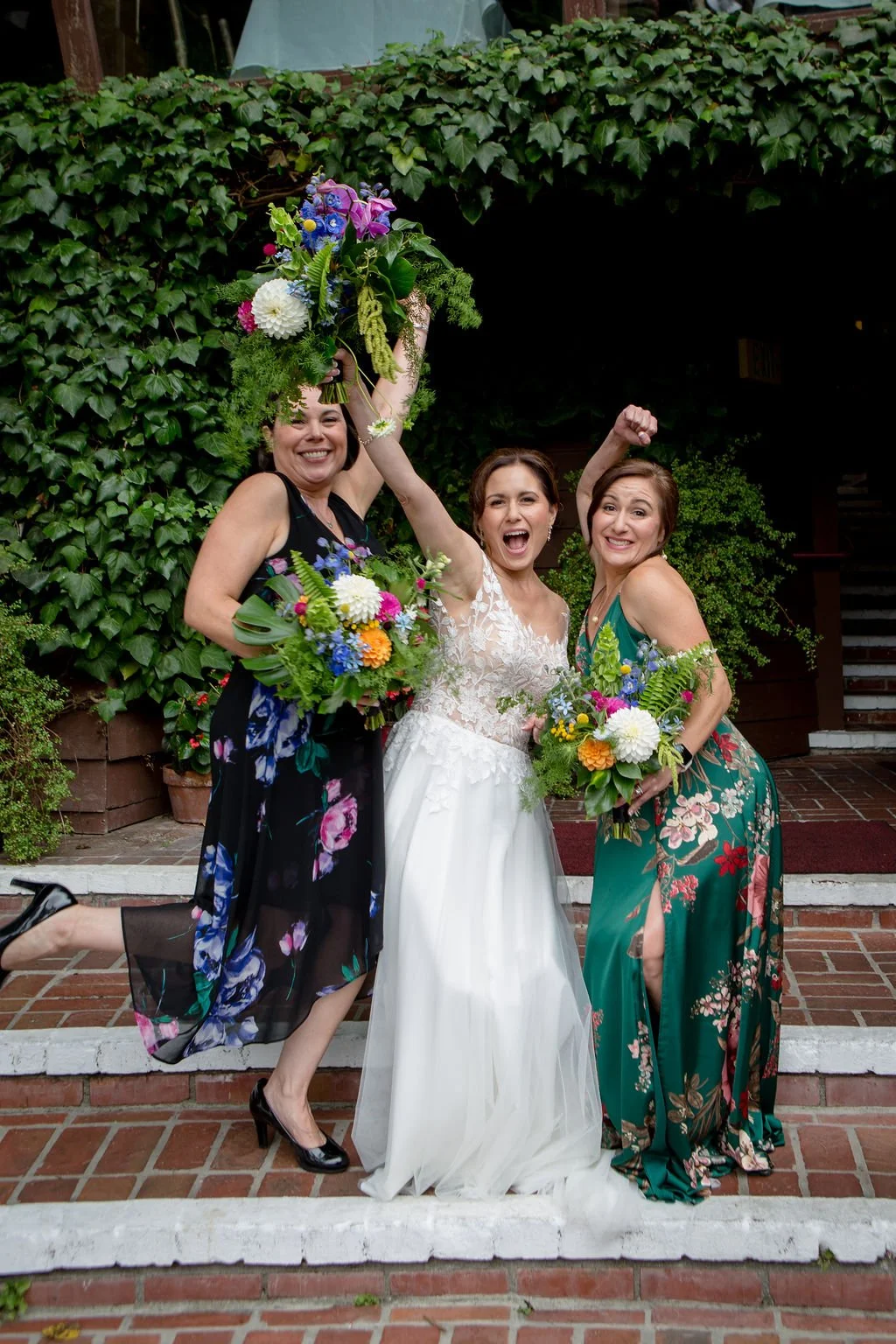 Three women in formal dresses joyfully posing with colorful bouquets at an outdoor event.