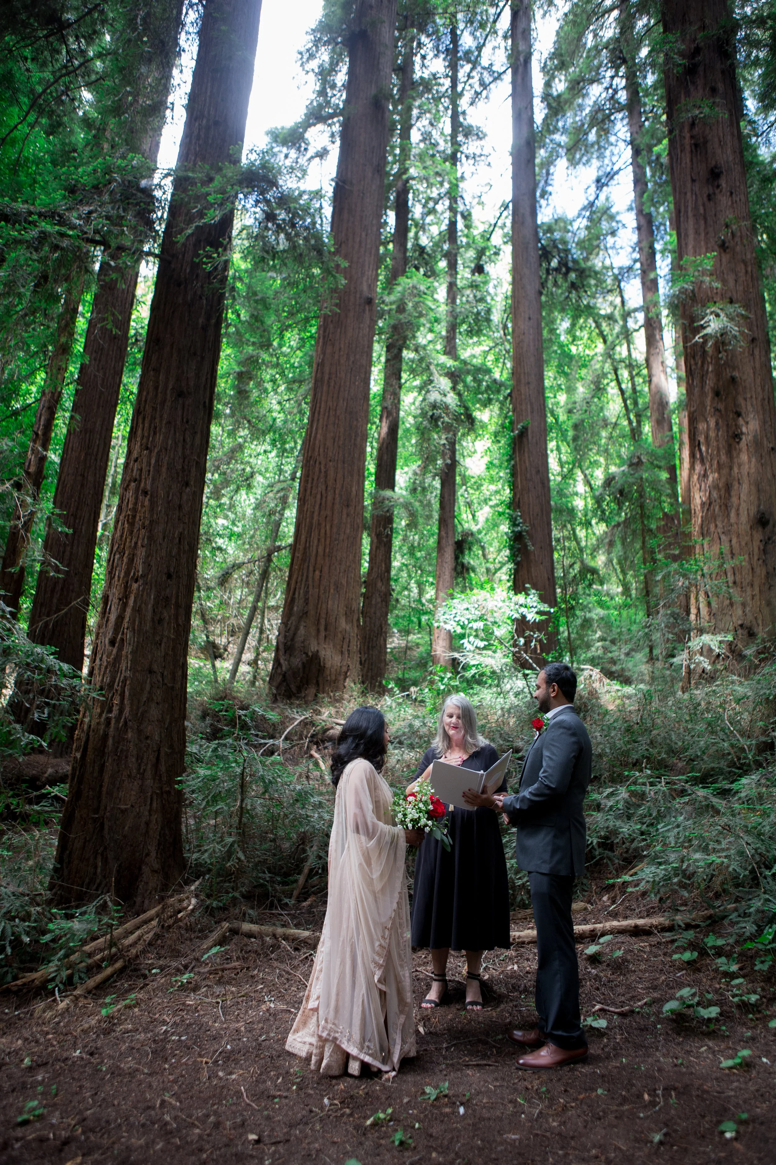 A couple getting married in a forest setting, surrounded by tall trees. A woman officiating the ceremony stands facing them, holding papers. The bride is wearing a white dress and holding flowers, while the groom is in a suit.