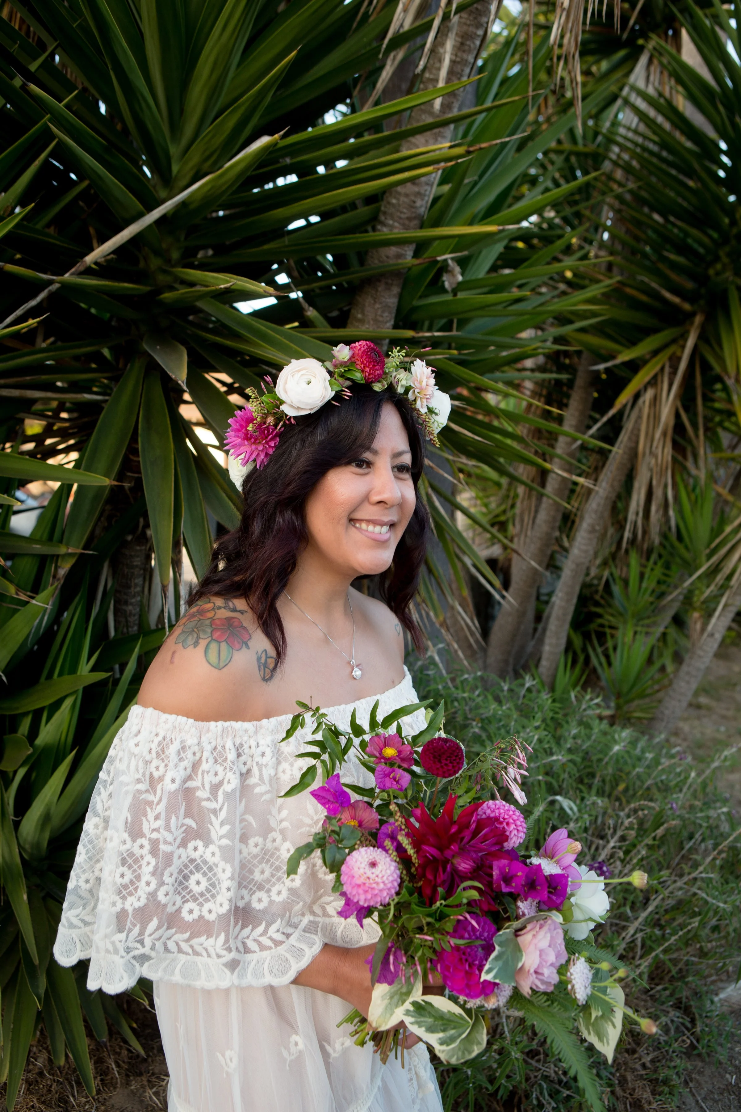 Woman in white lace dress with floral crown holding bouquet, standing outdoors with tropical plants.