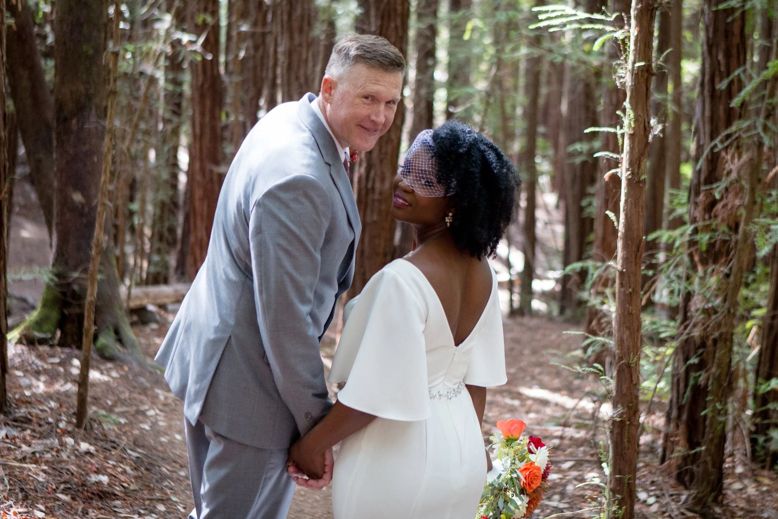 Bride and groom holding hands in a redwood forest, with the groom in a gray suit and the bride in a white dress carrying a colorful bouquet.