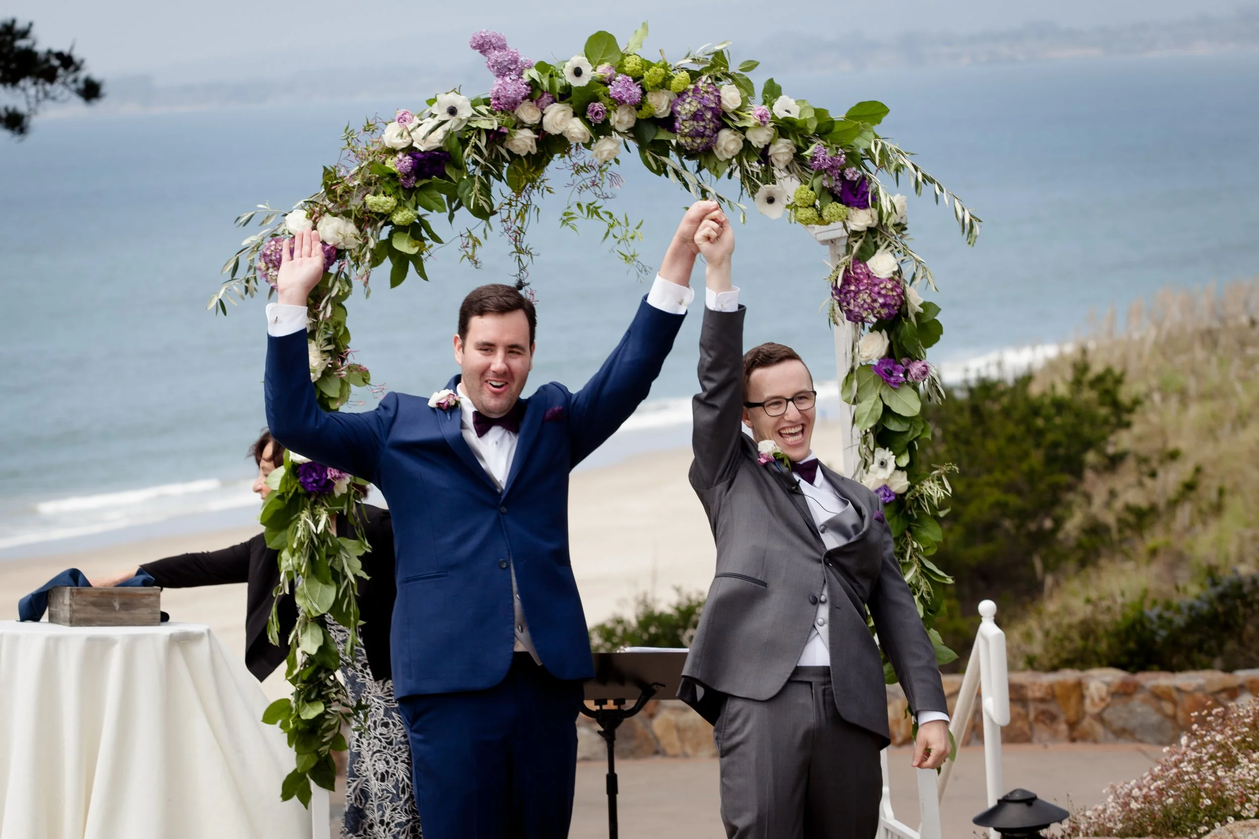 Two grooms celebrating under a floral arch at a beach wedding ceremony.