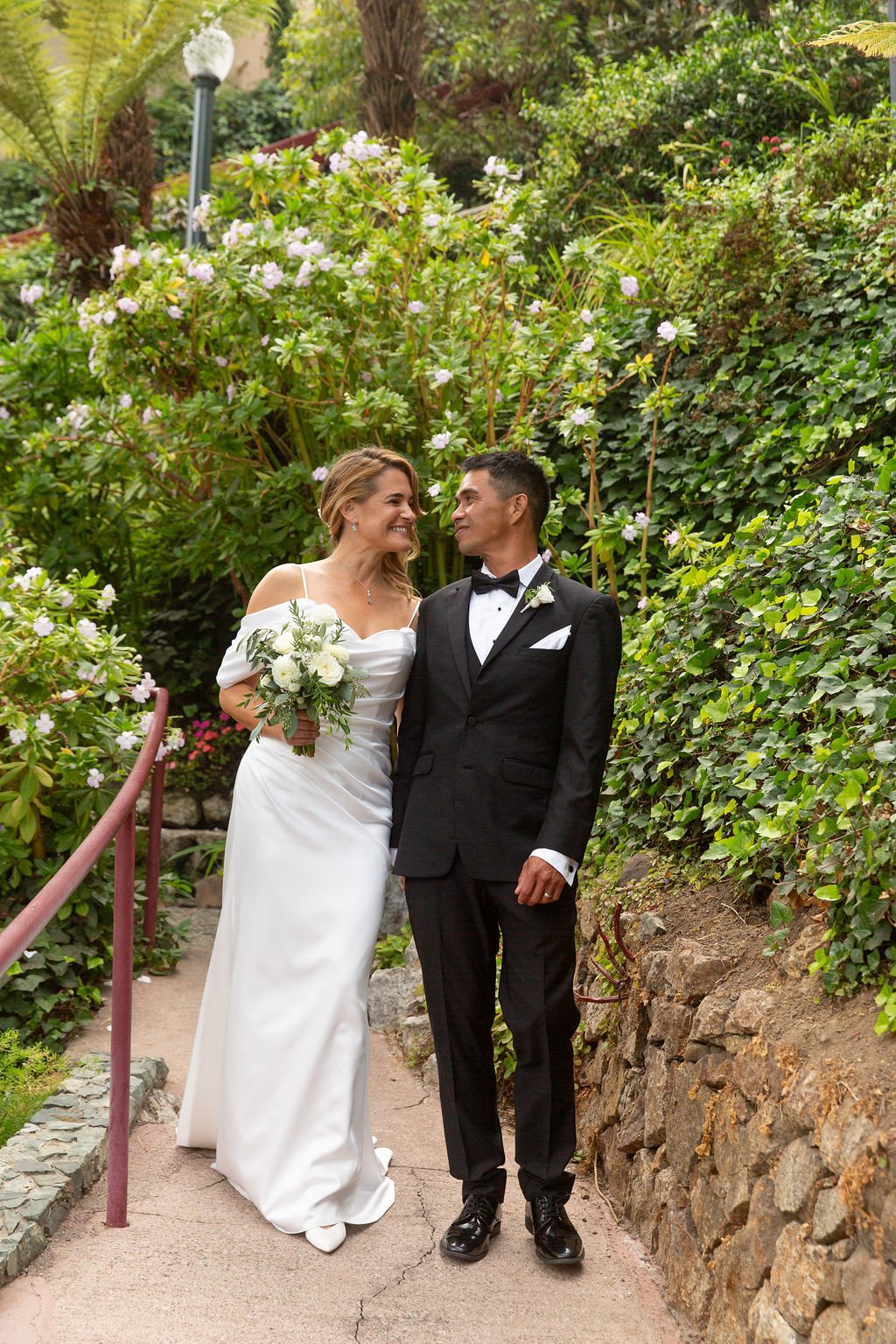 Bride and groom standing on a garden path with greenery and flowers, bride wearing a white dress holding a bouquet, groom in a black tuxedo.
