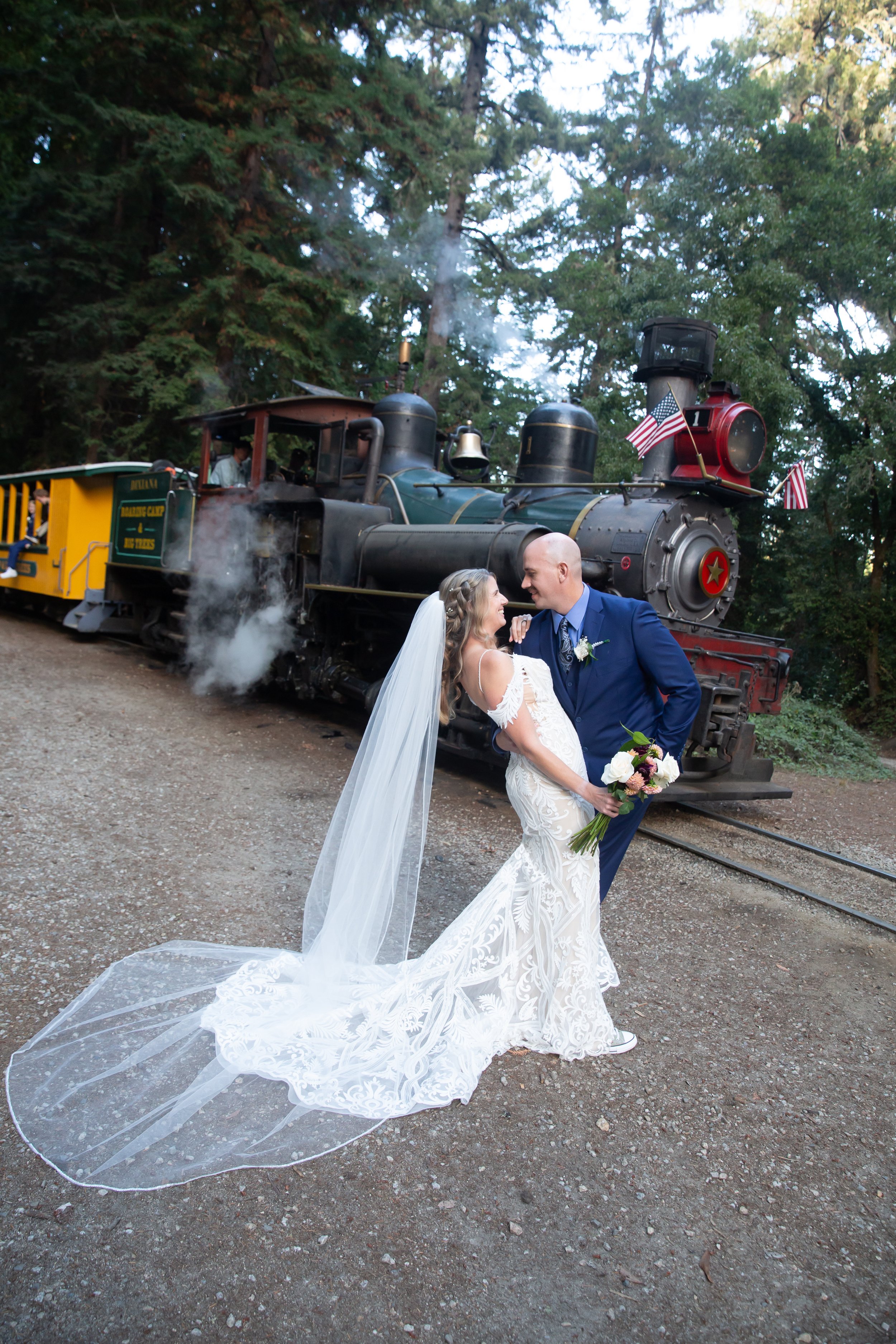 Bride and groom posing in front of a vintage steam train, surrounded by trees. The bride is wearing a lace wedding dress with a long veil and holding a bouquet, while the groom is in a blue suit. American flags adorn the front of the train.