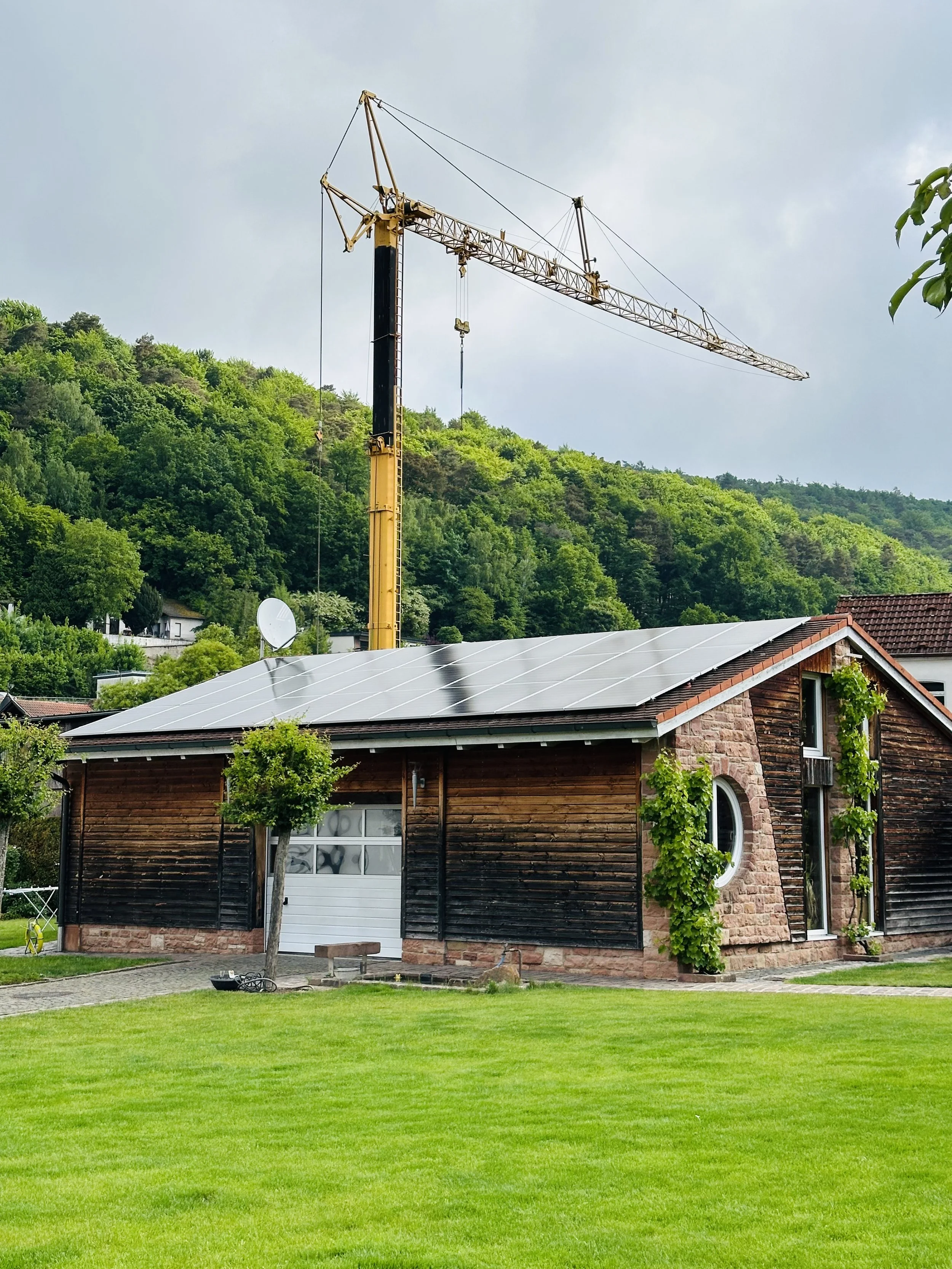 Ein Haus mit Solarpanelen auf dem Dach und einem Baukran im Hintergrund, umgeben von grüner Natur.