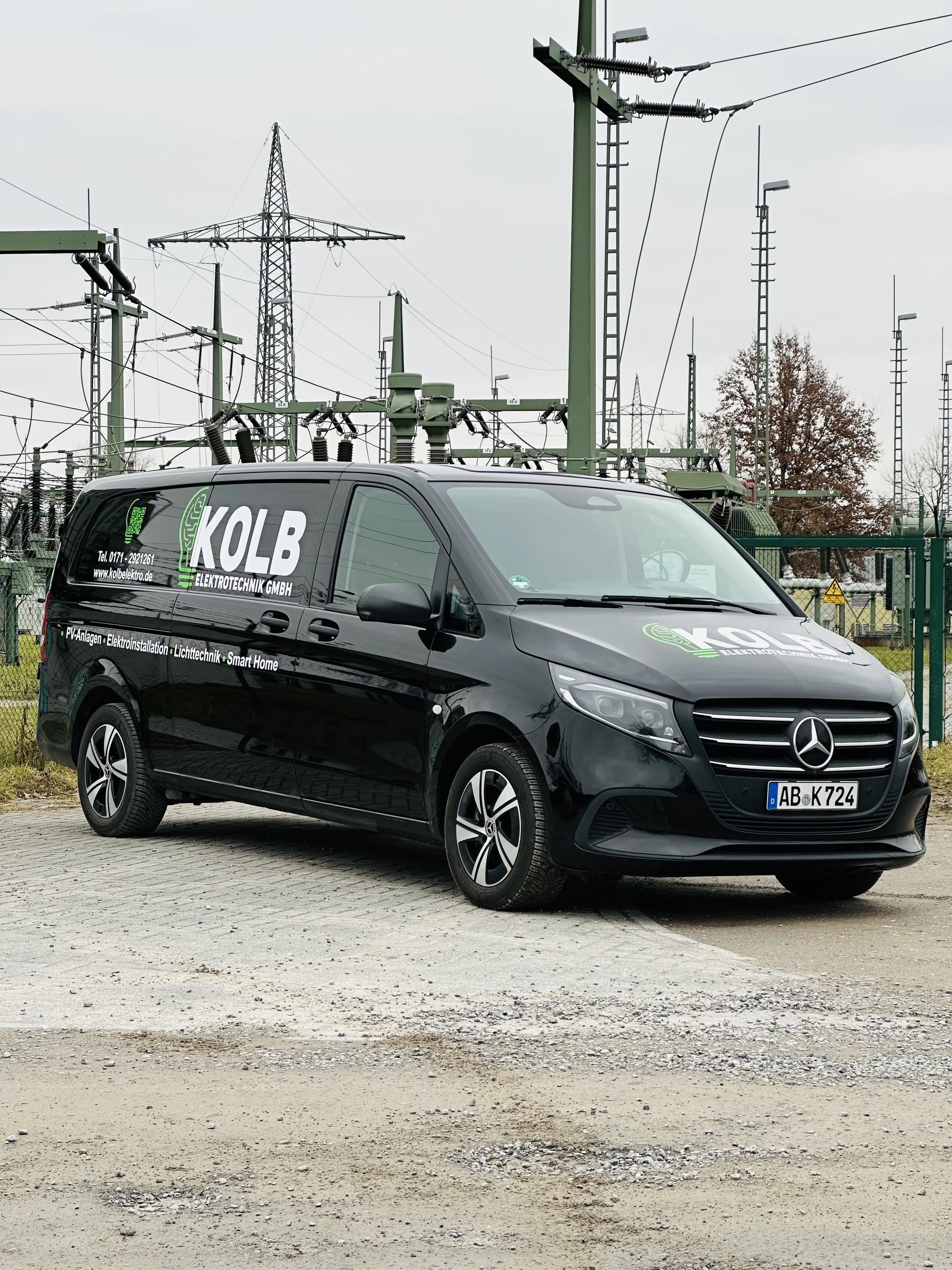 Schwarzer Mercedes-Benz-Van mit Firmenbeschriftung für Elektrotechnik, parkend auf einer unbefestigten Fläche vor einem Umspannwerk mit Hochspannungsmasten und Kabeln.