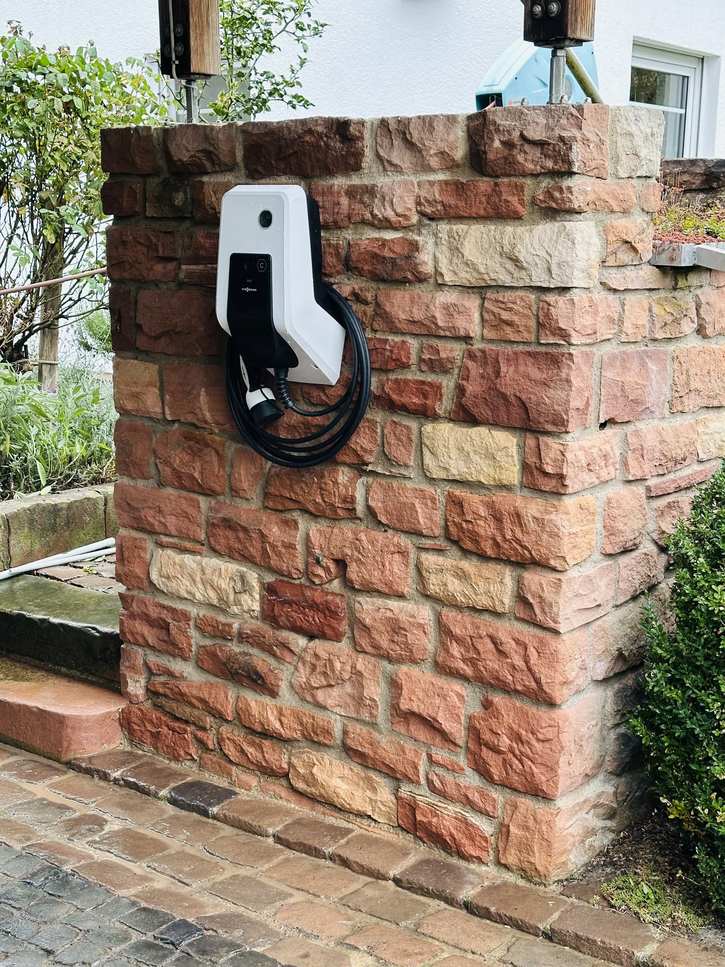 Ein Ladestation für Elektroautos an einer roten Ziegelmauer im Garten, umgeben von grünen Pflanzen.