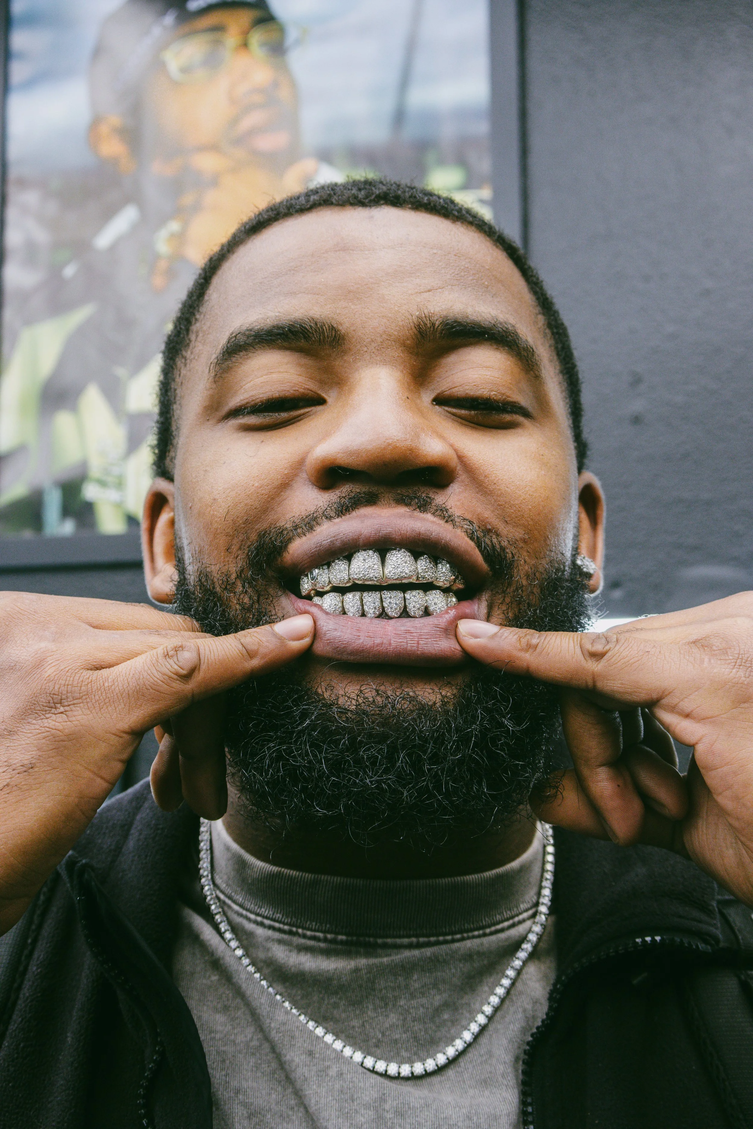 A man with a beard and closed eyes is smiling as someone points to his mouth, revealing a set of silver grillz on his teeth. There is a portrait of a person in sunglasses and a yellow jacket in the background.